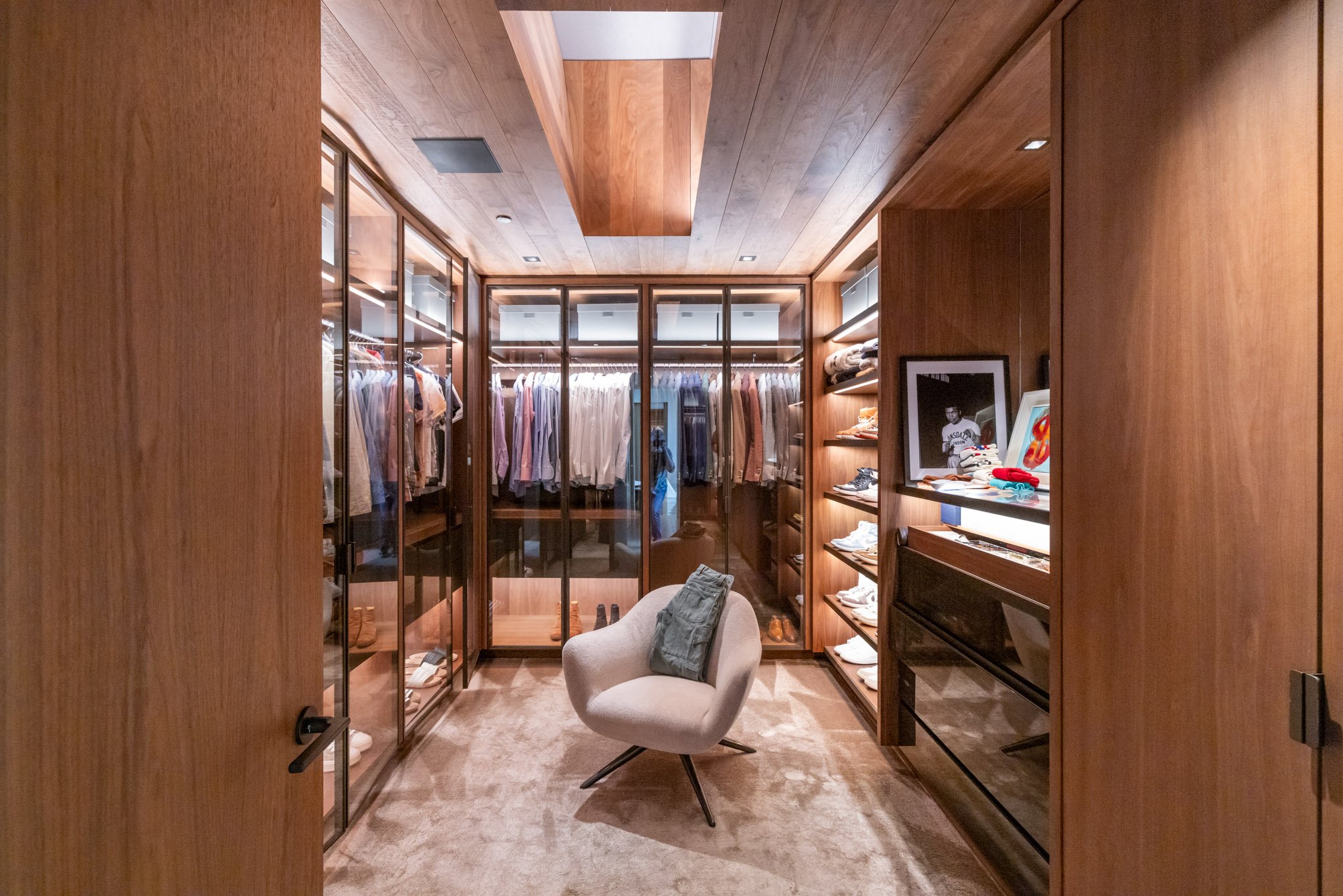 A walk-in closet with wooden walls, glass wardrobes filled with clothes, shelves with shoes, and a beige chair with a folded denim shirt on it.