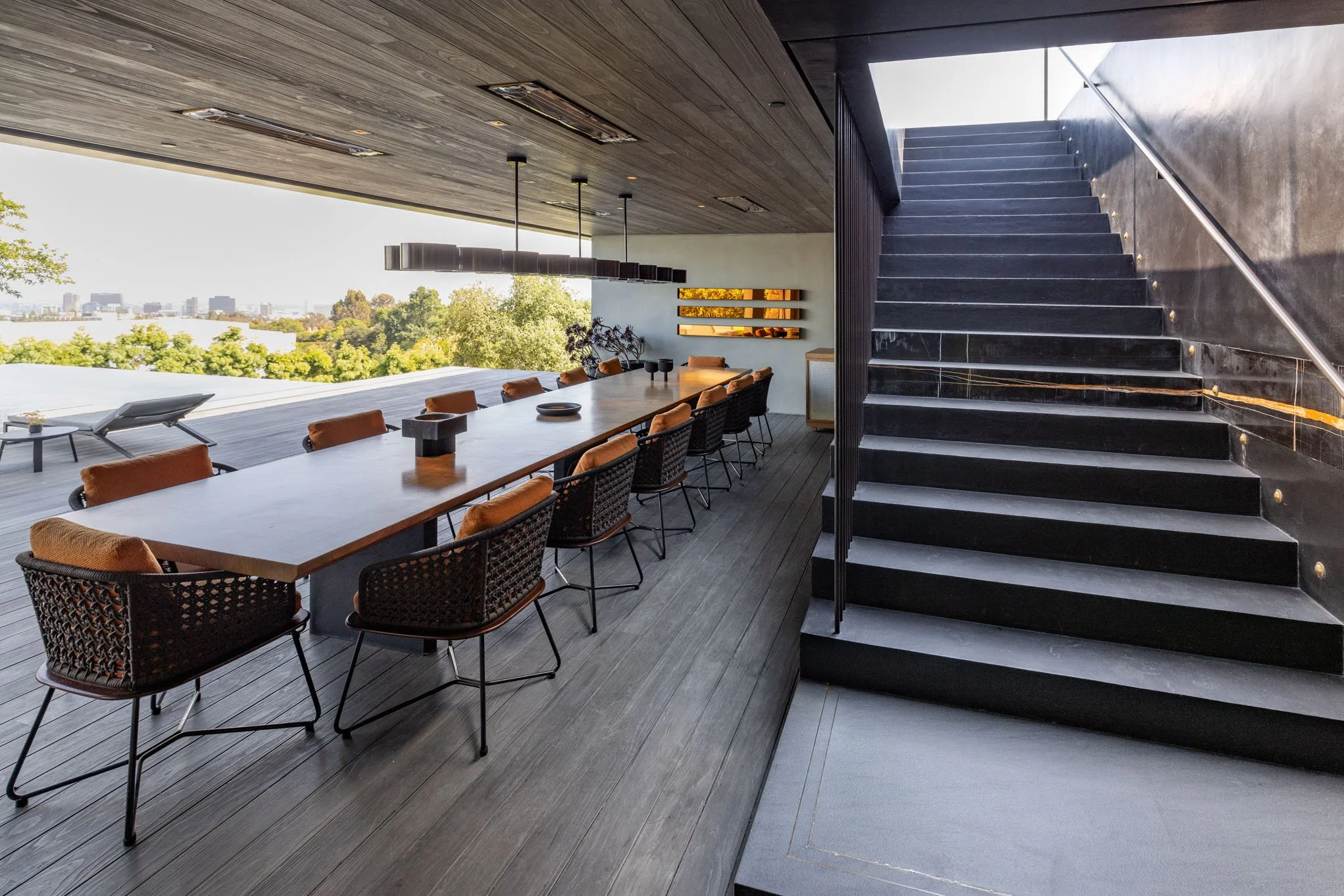Modern dining area with a long wooden table, ten chairs with woven backs and caramel-colored cushions, and a view of trees and the city skyline through an open wall. A staircase with black steps and a metallic handrail is on the right.