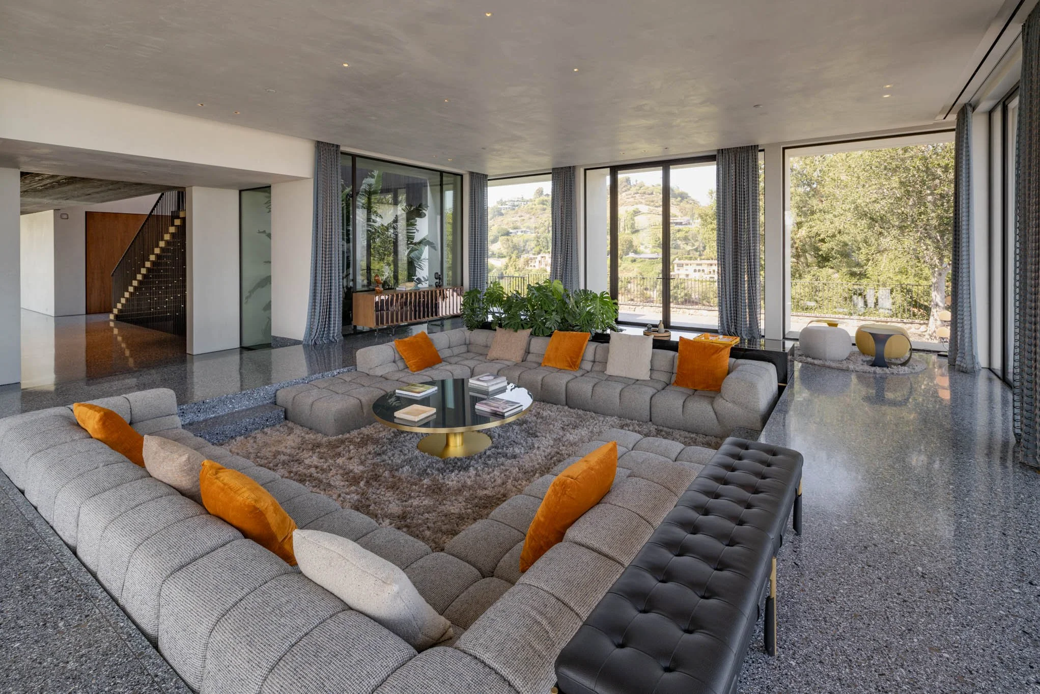 Modern living room with large sectional sofa, orange and neutral pillows, a round black coffee table, a textured area rug, and large windows with scenic outdoor views.