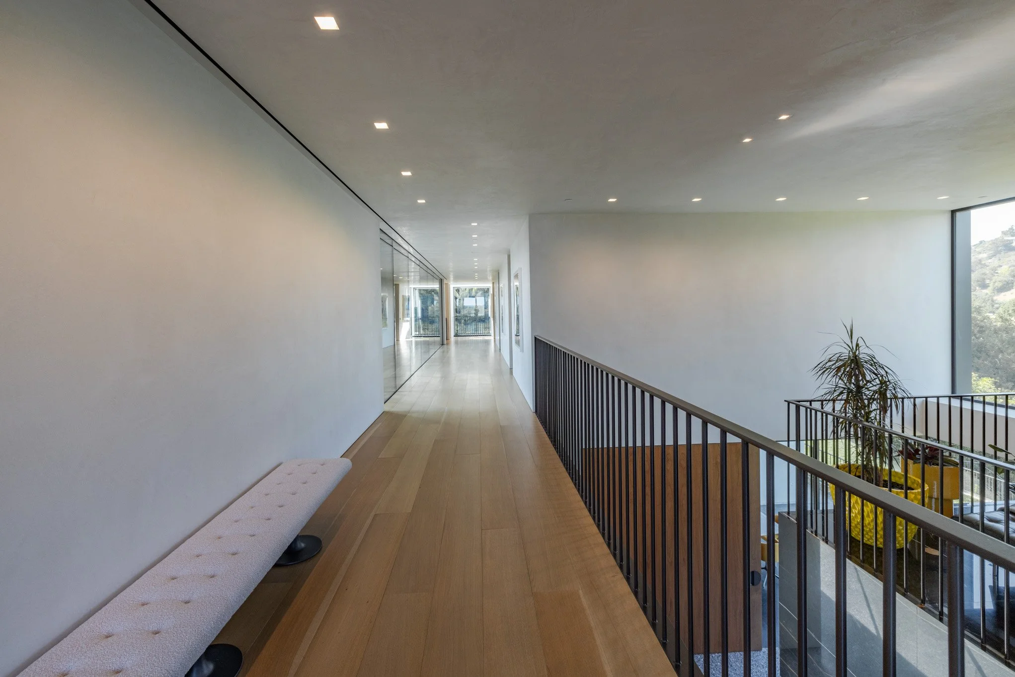 Empty modern corridor with wooden floor, white walls, ceiling lights, glass wall on the left, and large window on the right with a view of green trees and a potted plant.