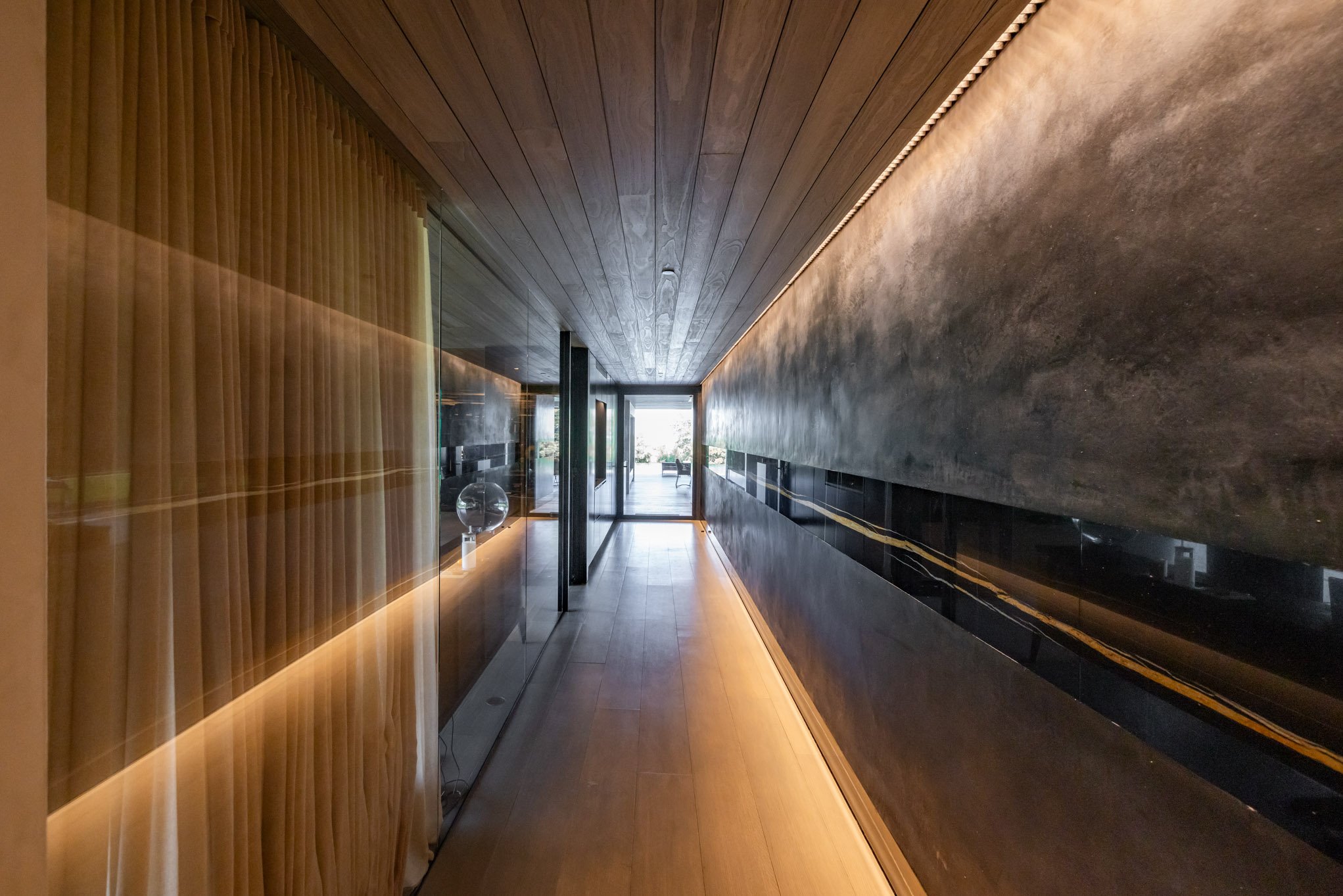 A modern, narrow hallway with wooden ceiling and floor, black and gray textured walls, glass panels on the left, and a bright space at the end with outdoor furniture and trees.