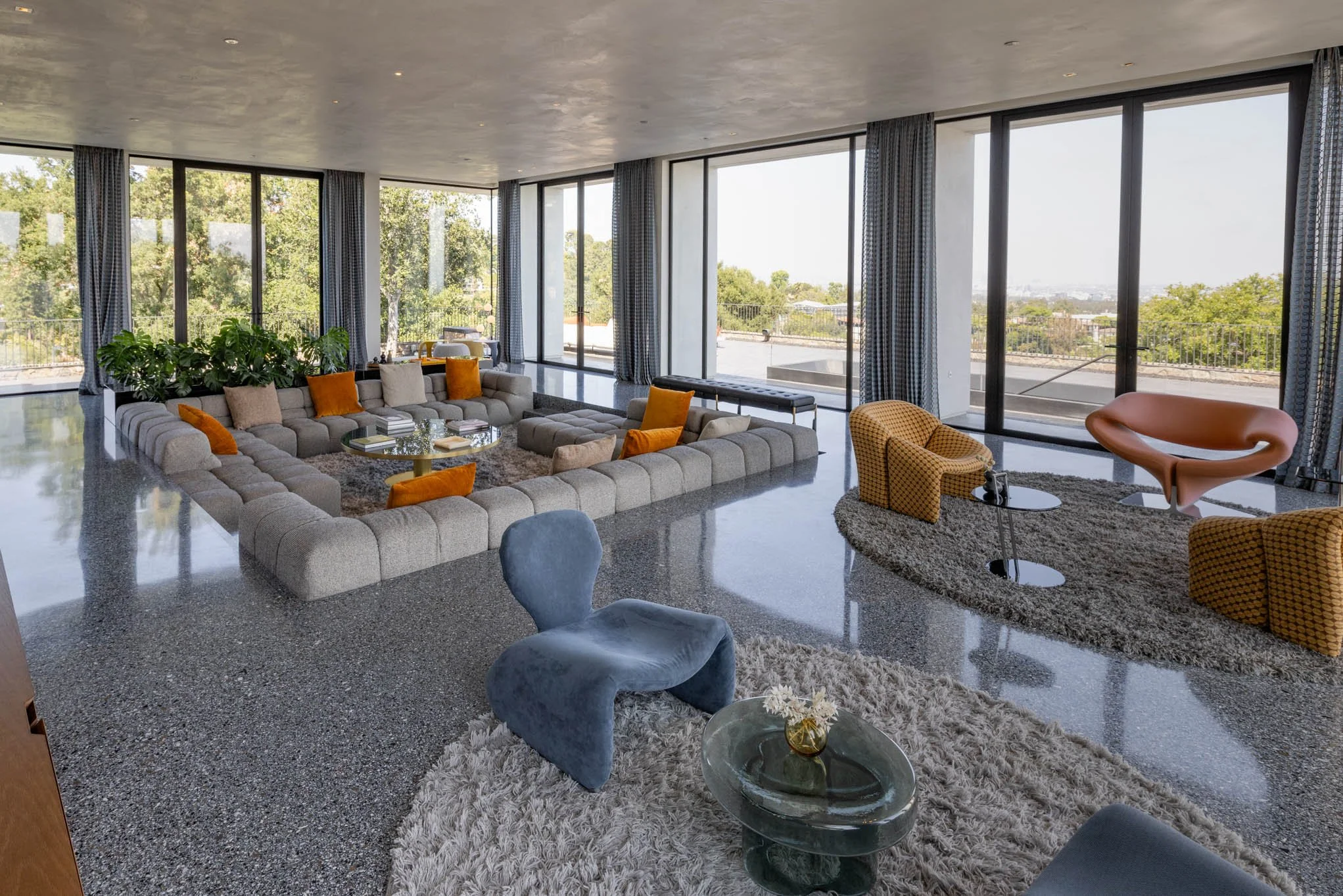 Modern living room with large windows, beige sectional sofa with orange and neutral pillows, eclectic chairs, glass coffee tables, green plants, and view of greenery outside.