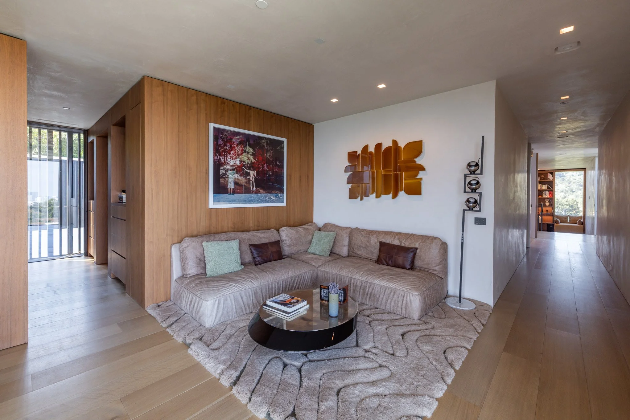 Modern living room with beige sectional sofa, decorative pillows, a round black coffee table, wooden wall panel, abstract wall art, and a textured rug.