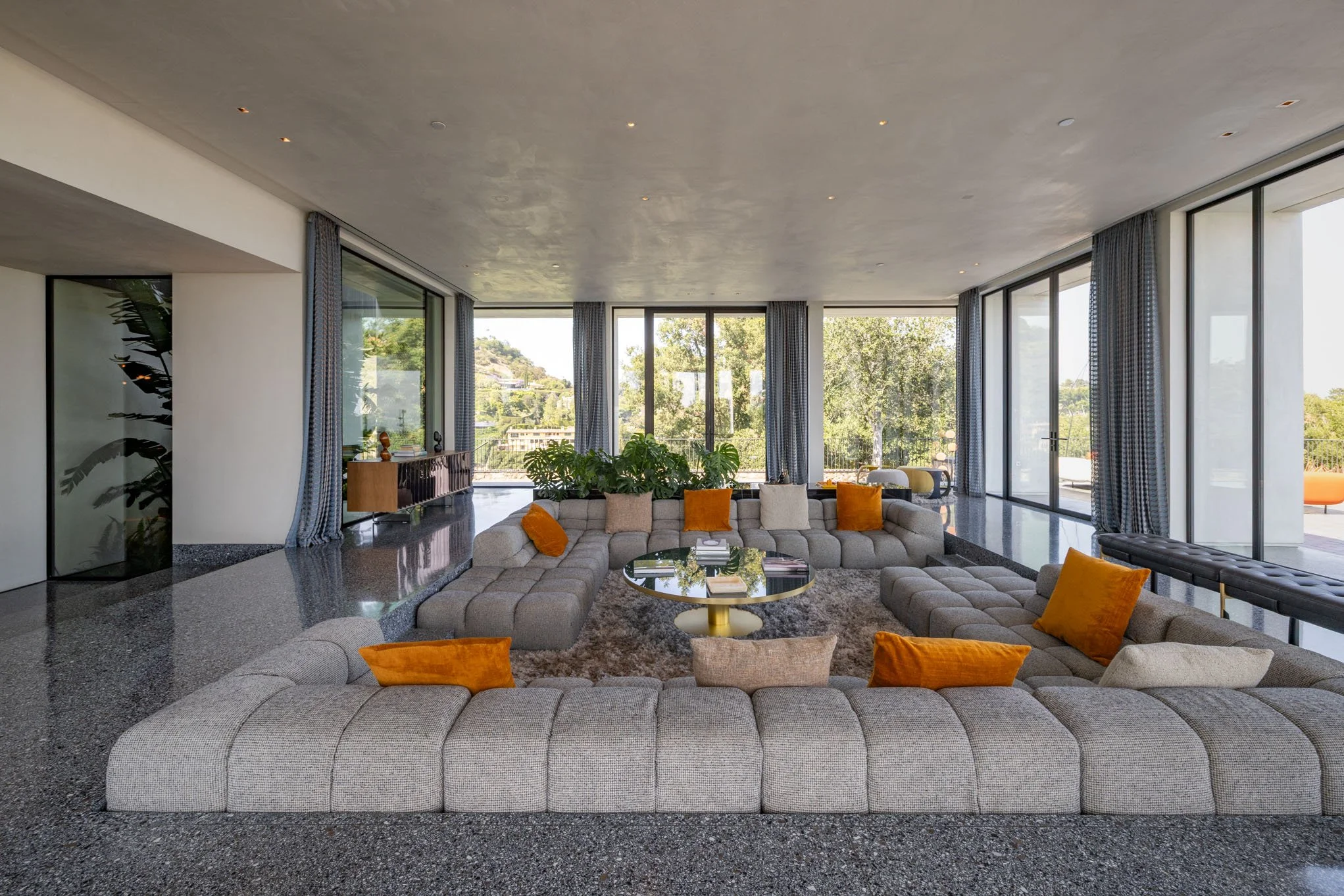 Spacious modern living room with large windows, sectional gray sofa with orange and beige pillows, glass-top coffee table, indoor plants, and curtains, overlooking a lush outdoor landscape.
