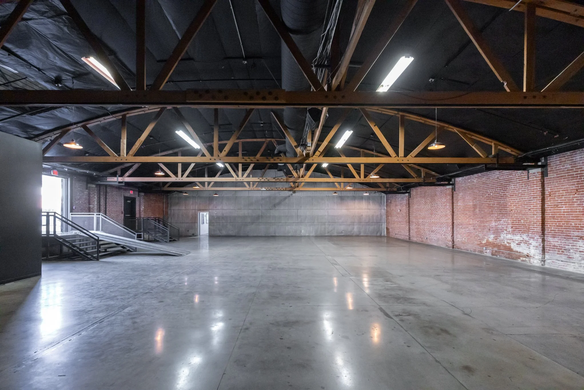 Empty industrial-style event space with polished concrete floors, brick walls, exposed wooden beams, and fluorescent and hanging lights.