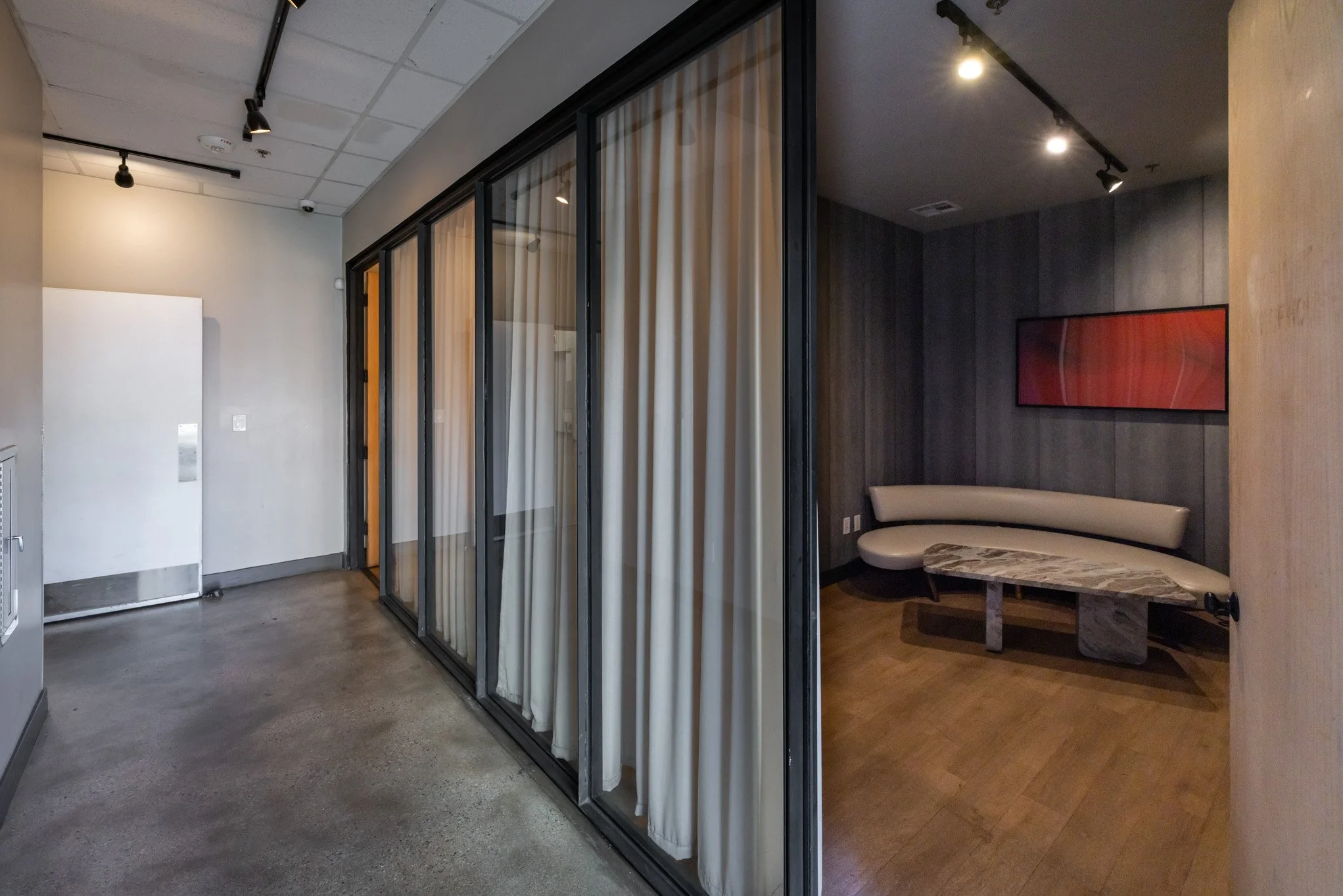 Interior view of a modern office lounge with a curved sofa, marble coffee table, dark wood paneled wall, adjacent glass-walled meeting room with beige curtains, and ceiling track lighting.