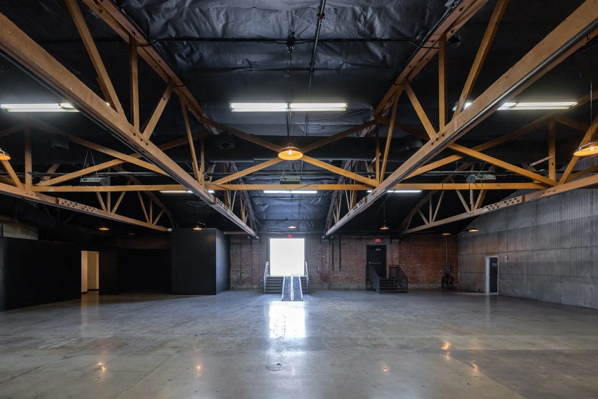 Empty warehouse or industrial space with exposed wooden beams, overhead lighting, and a concrete floor, illuminated by a large doorway at the back.