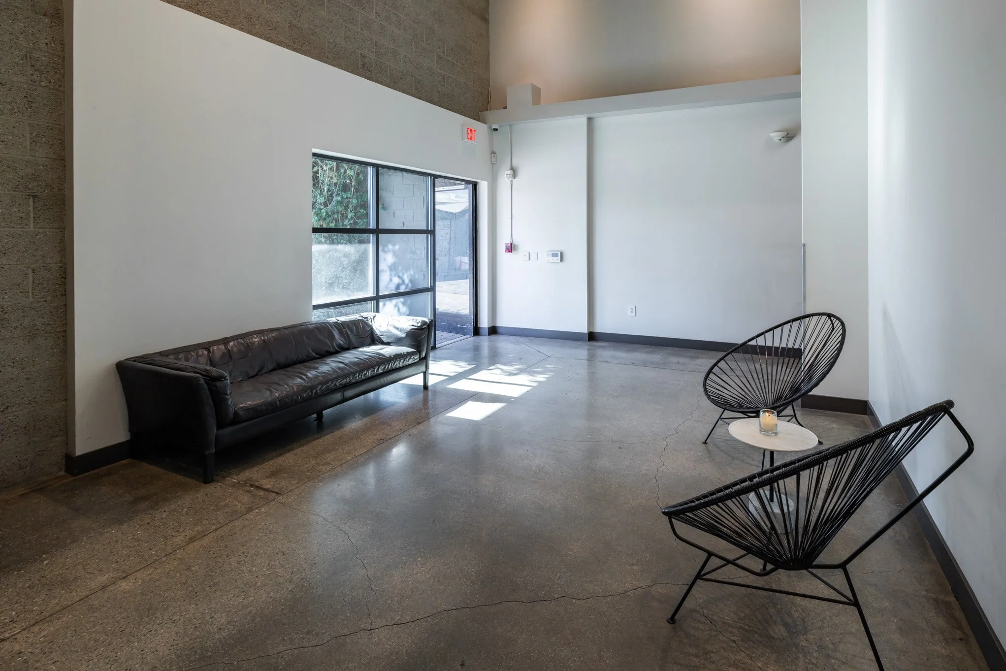 Minimalist waiting area with a black leather sofa and two black wire chairs with a small round table in between, holding a candle, inside a modern building with concrete floor and large window.