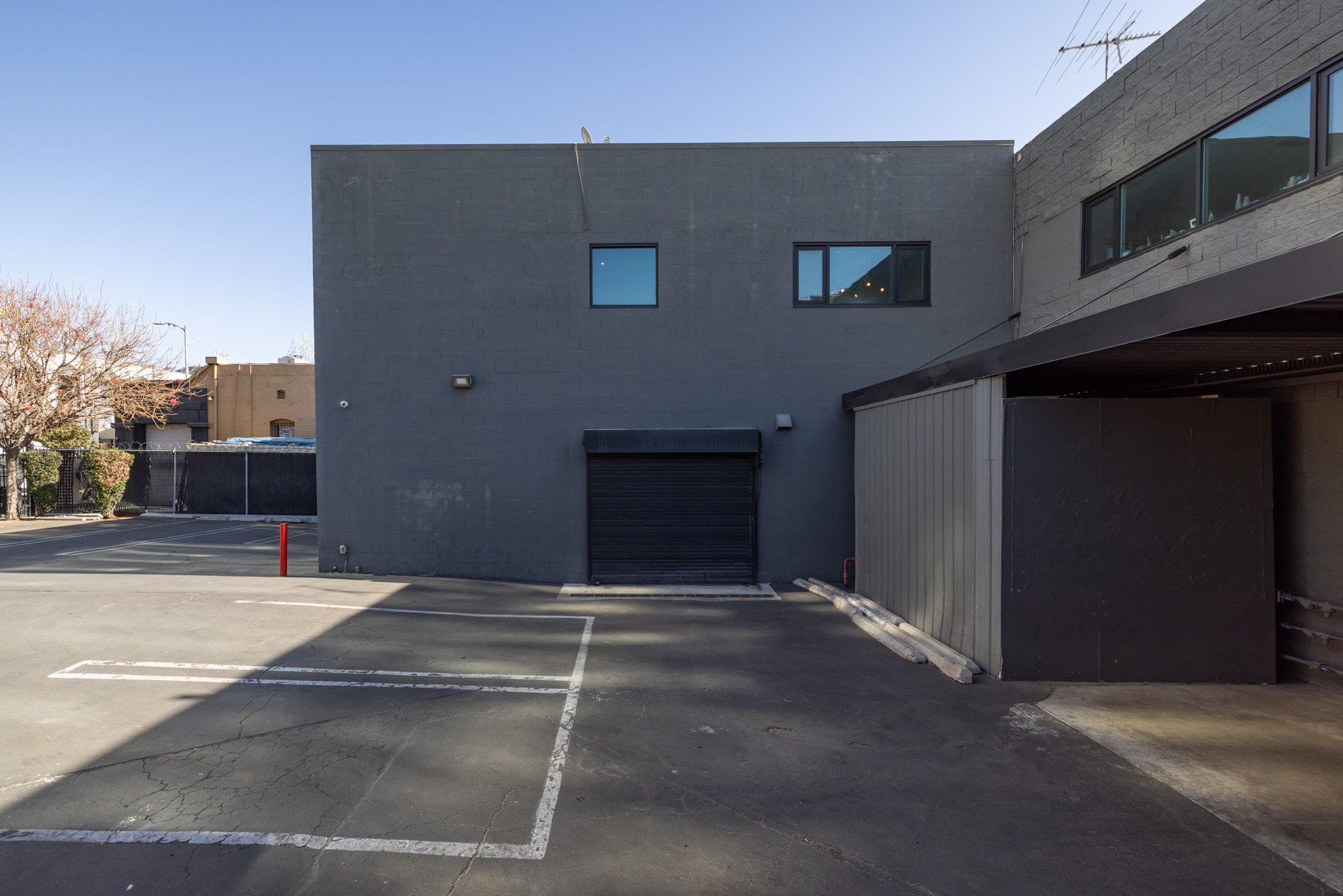Empty parking lot with marked spaces in front of a gray industrial building, with a closed black roll-up door and a small window, under a clear blue sky.