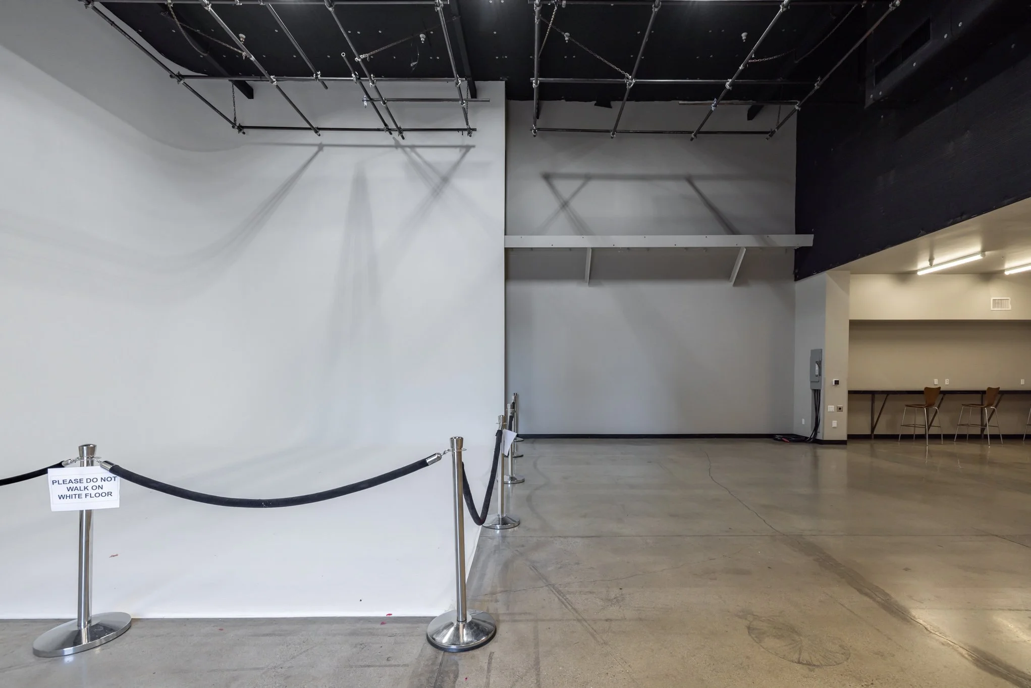Empty indoor space with a white wall, black ceiling, and concrete floor, sectioned off with a black rope barrier and a sign that reads 'Please do not walk on white floor.' In the background, there are two chairs and a counter with a wall-mounted phon
