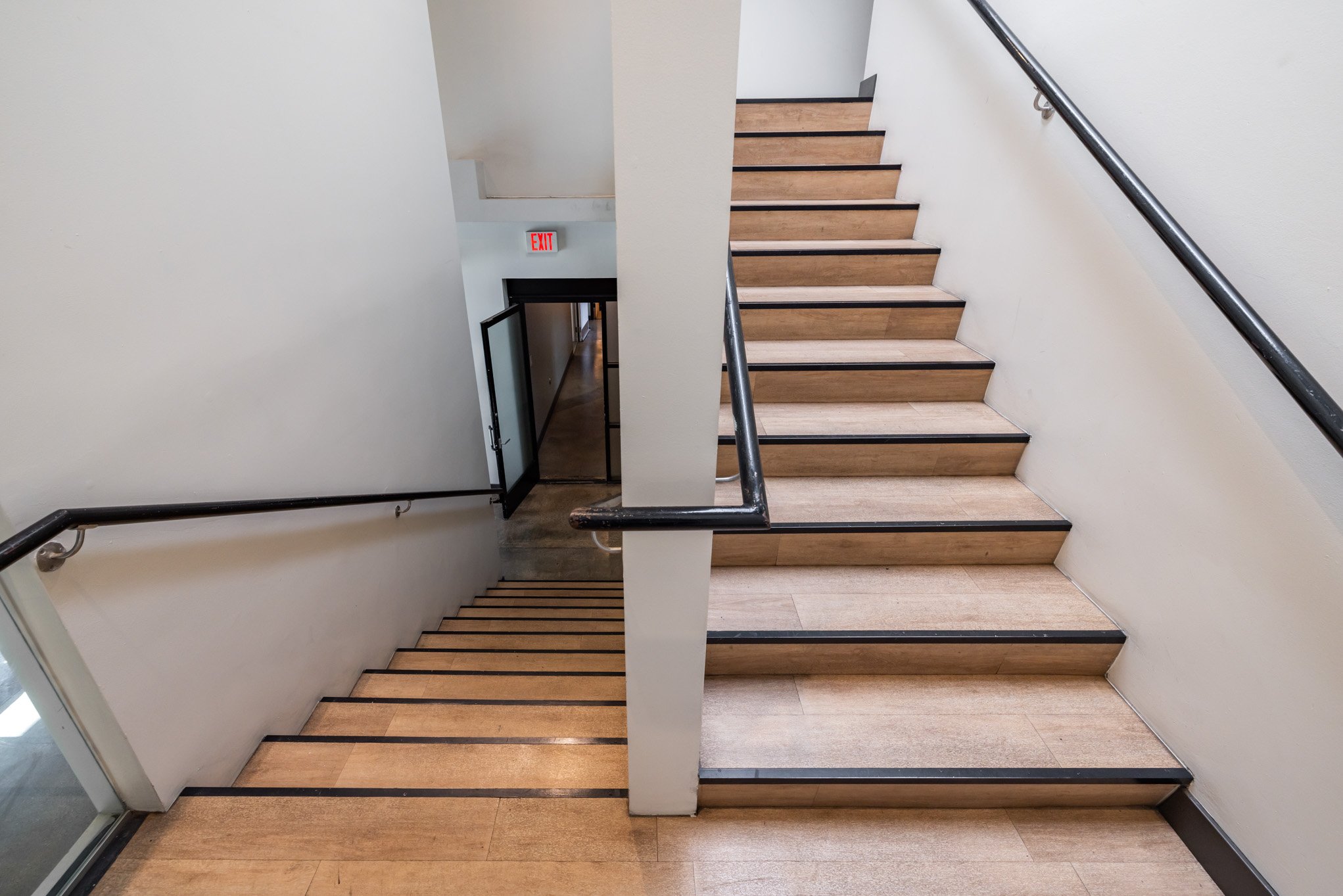 A staircase with wooden steps, black edge trim, and a black metal handrail leading to a corridor in a building, with an exit door and an emergency exit sign overhead.