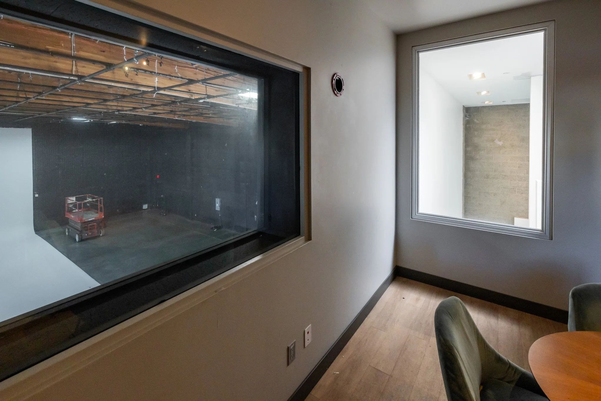 Empty room with wood flooring, beige walls, and two large windows looking into an adjacent room under construction, with visible drywall and ceiling lights.