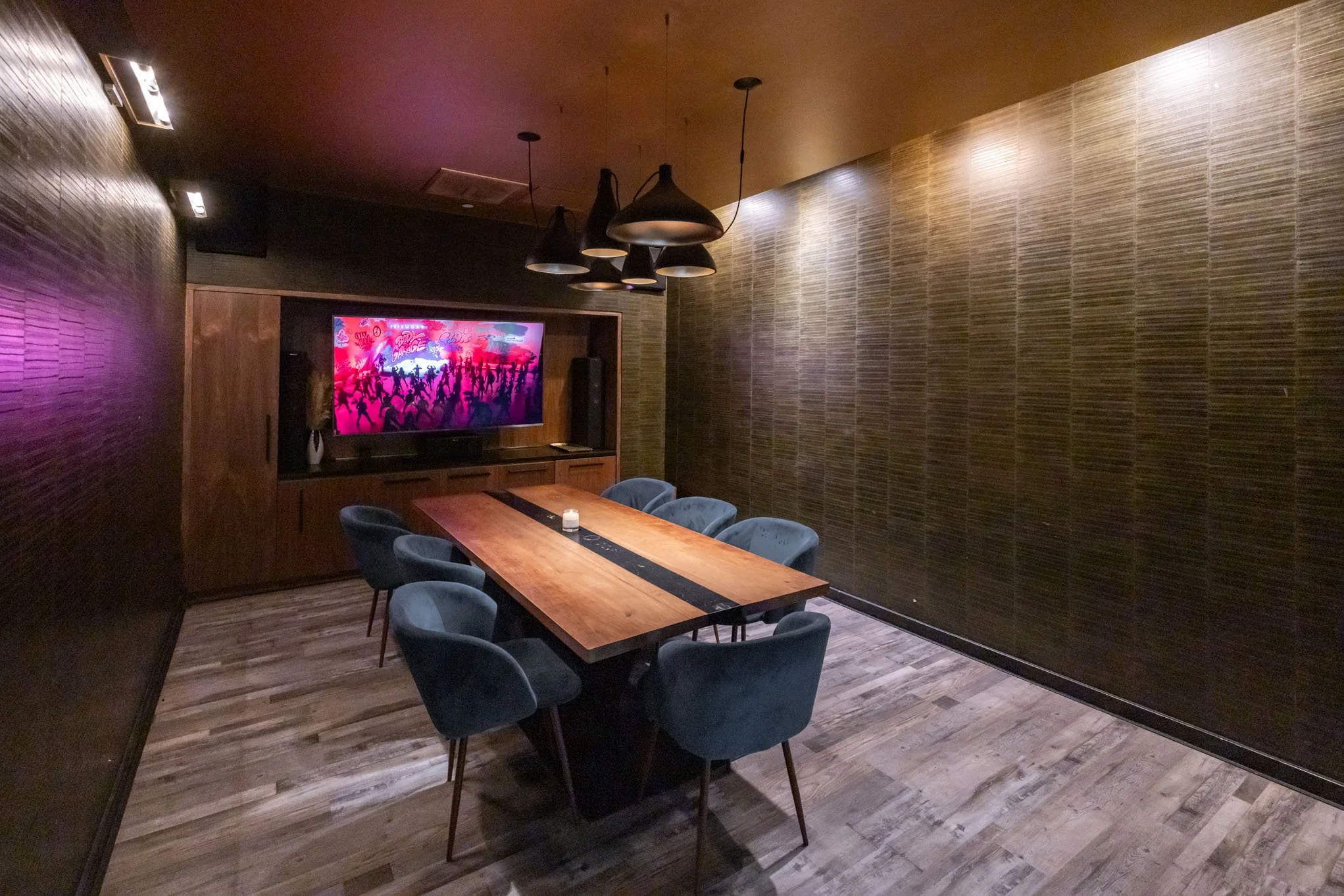 A modern private room with a wooden dining table surrounded by six blue upholstered chairs, a wall-mounted flat-screen TV displaying a dance scene, and dark wood paneled walls with horizontal textured patterns. Overhead black pendant lights hang from