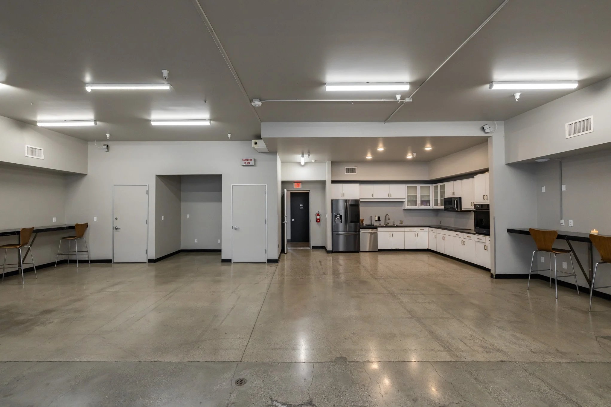 Empty communal kitchen and seating area with white cabinets, stainless steel refrigerator, black countertops, and bar stools along the walls.
