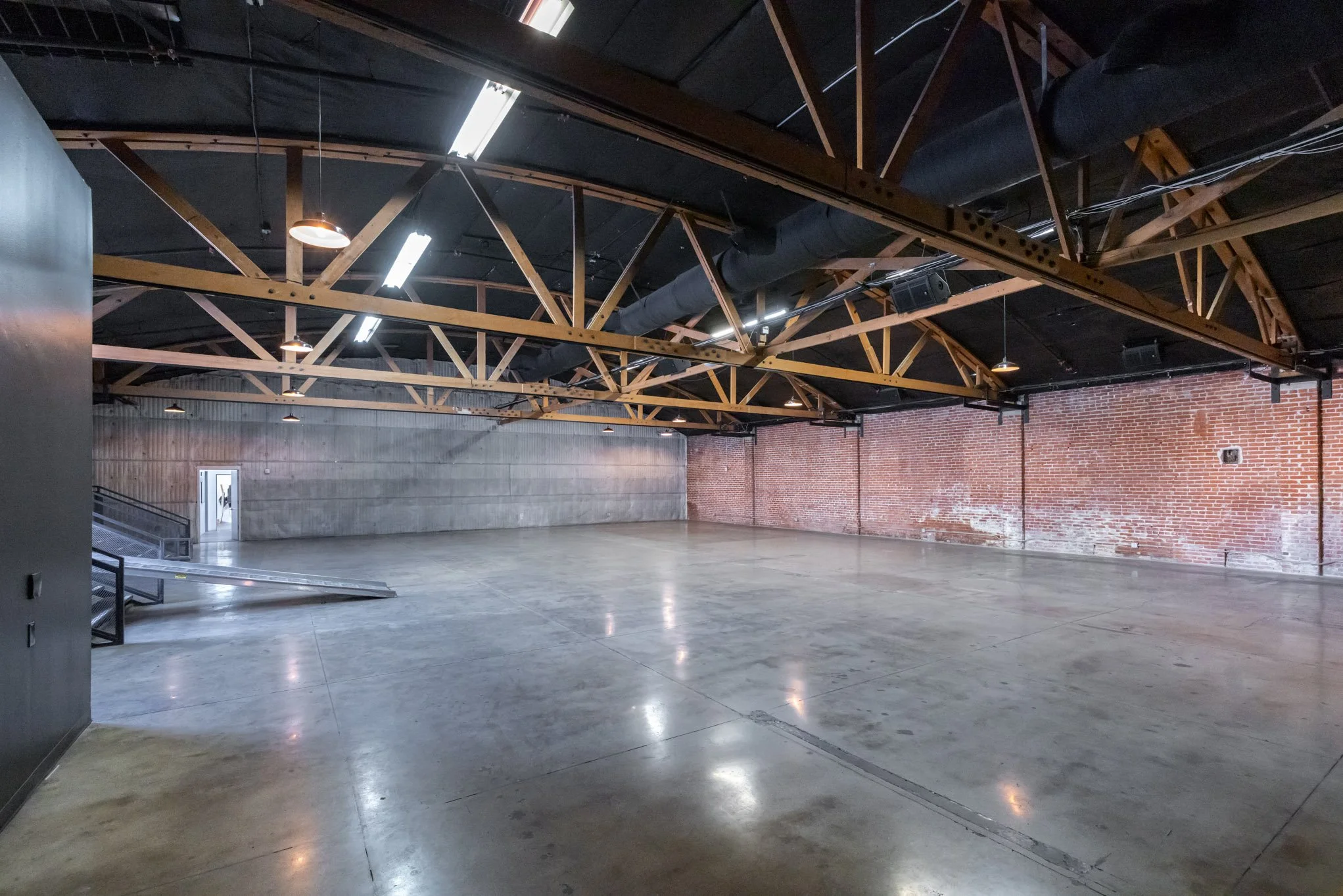 Empty industrial event space with polished concrete floors, exposed brick and concrete walls, and a ceiling with wooden beams and black ductwork, illuminated by fluorescent lights.