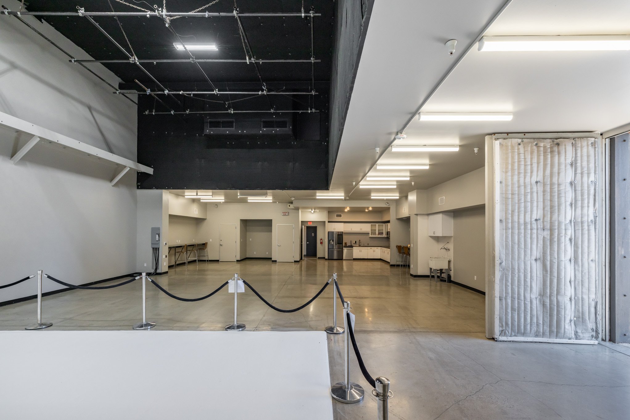 Empty event space with a black suspended stage, white walls, fluorescent lighting, kitchen area, and a quilted partition door on the right.