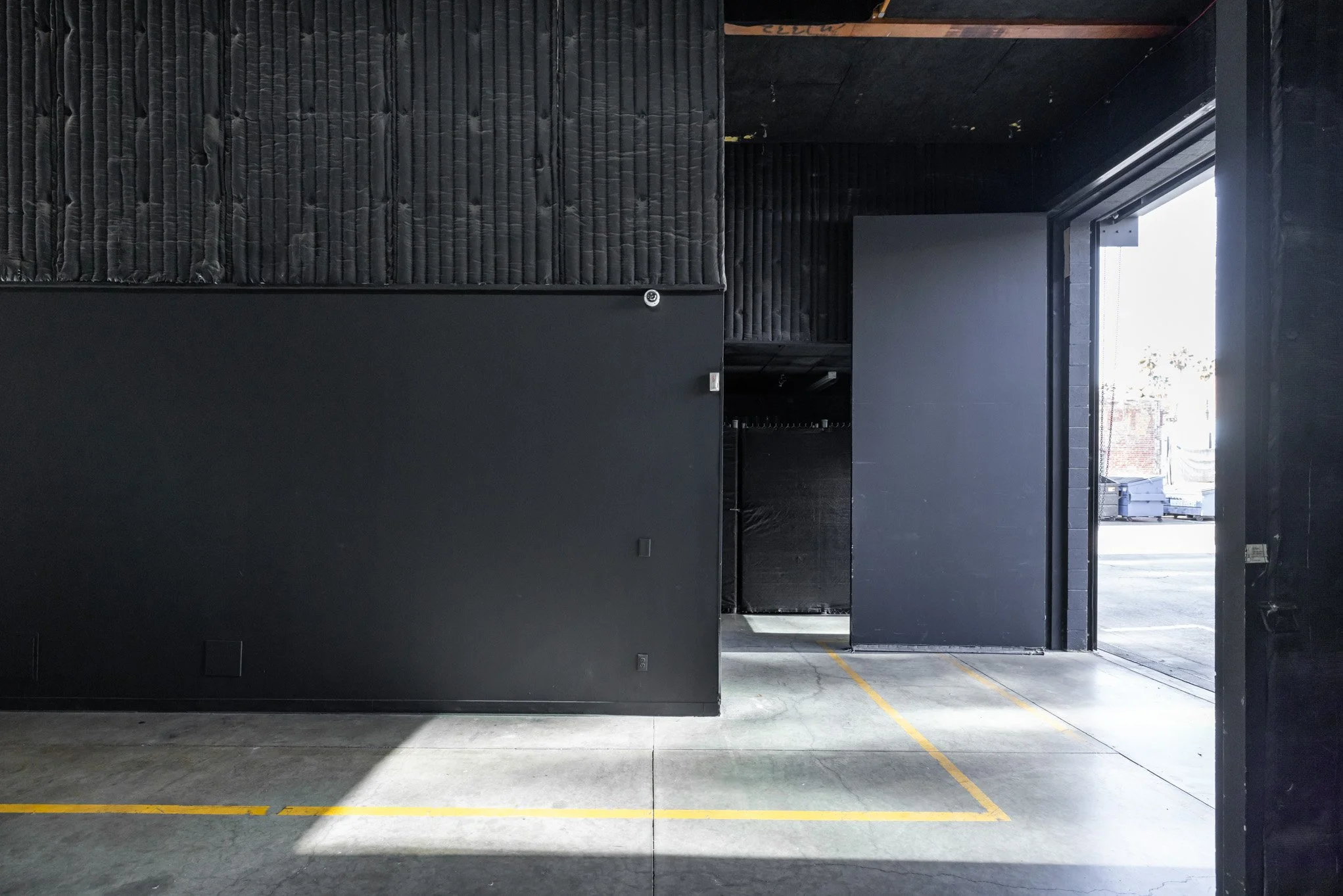 Empty parking space with yellow lines, open garage door showing outside view, black walls and ceiling, concrete floor.