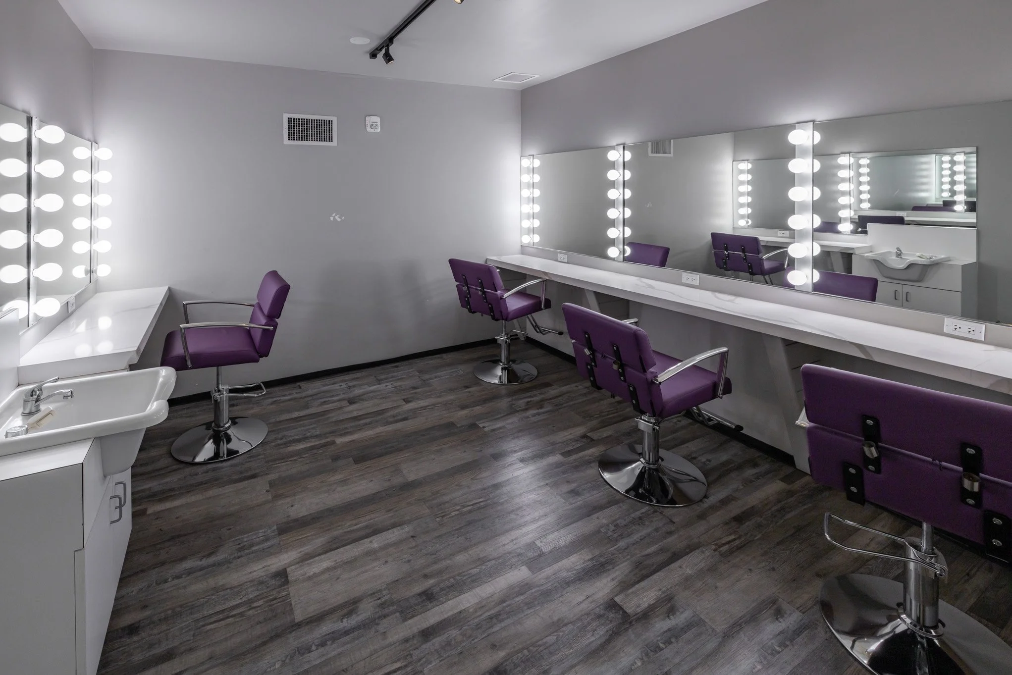Empty makeup or styling room with purple chairs, large mirrors with lights, a white countertop, and a small sink, in a modern salon or studio.
