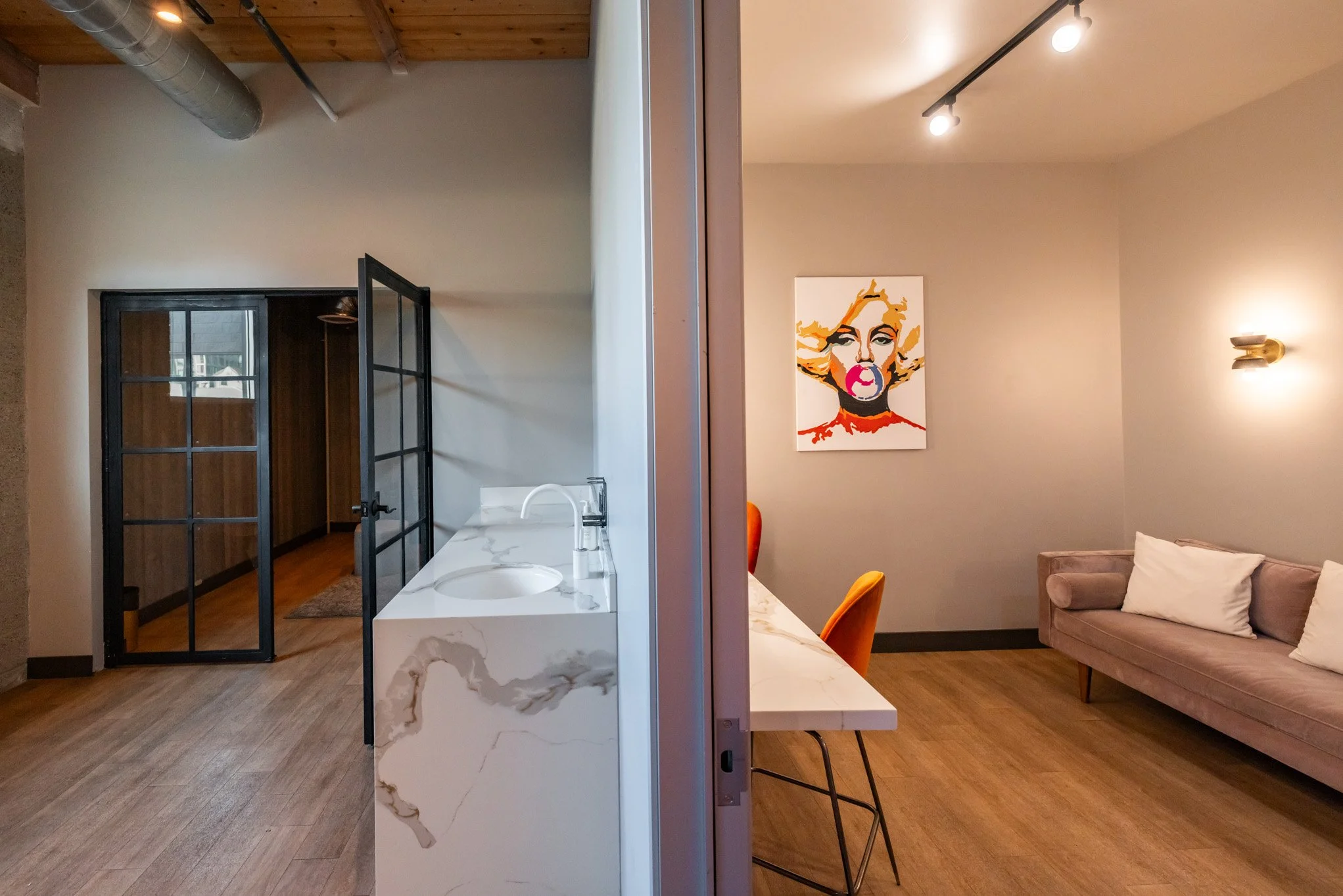 A split view of two rooms: the left side shows a minimalist kitchenette with a white marble countertop, a sink, and black framed glass doors leading to a hallway. The right side features a cozy living or dining area with a beige couch, white pillows,