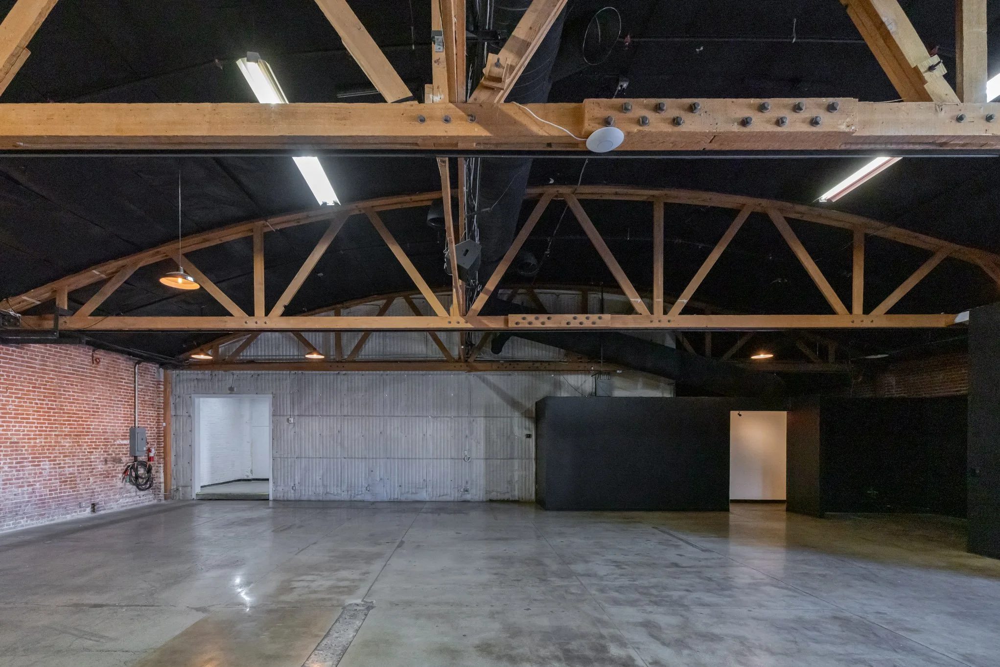 Empty industrial-style event space with concrete floors, brick and metal walls, exposed wooden beams, and black panels.