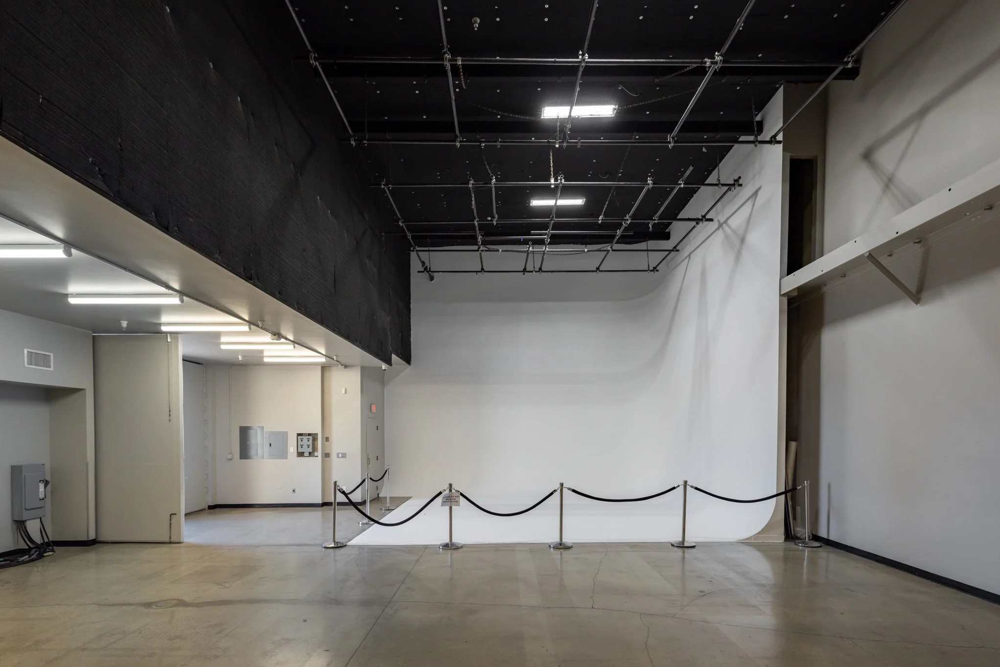 Empty museum or gallery space with a white wall, a black ceiling feature with pipes, and a roped-off area in the foreground.
