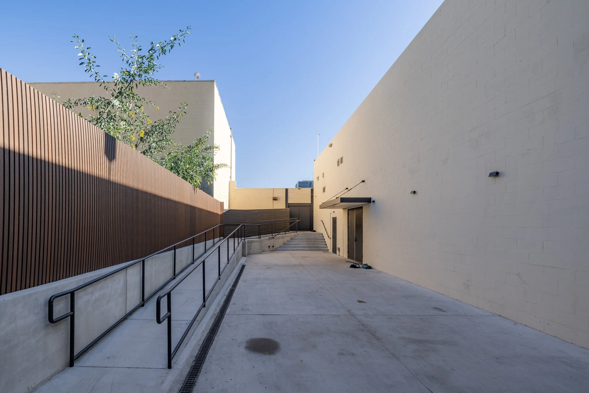 Empty outdoor pathway with a brown wooden fence on the left, beige walls on the right, stairs at the end, and a clear blue sky overhead.