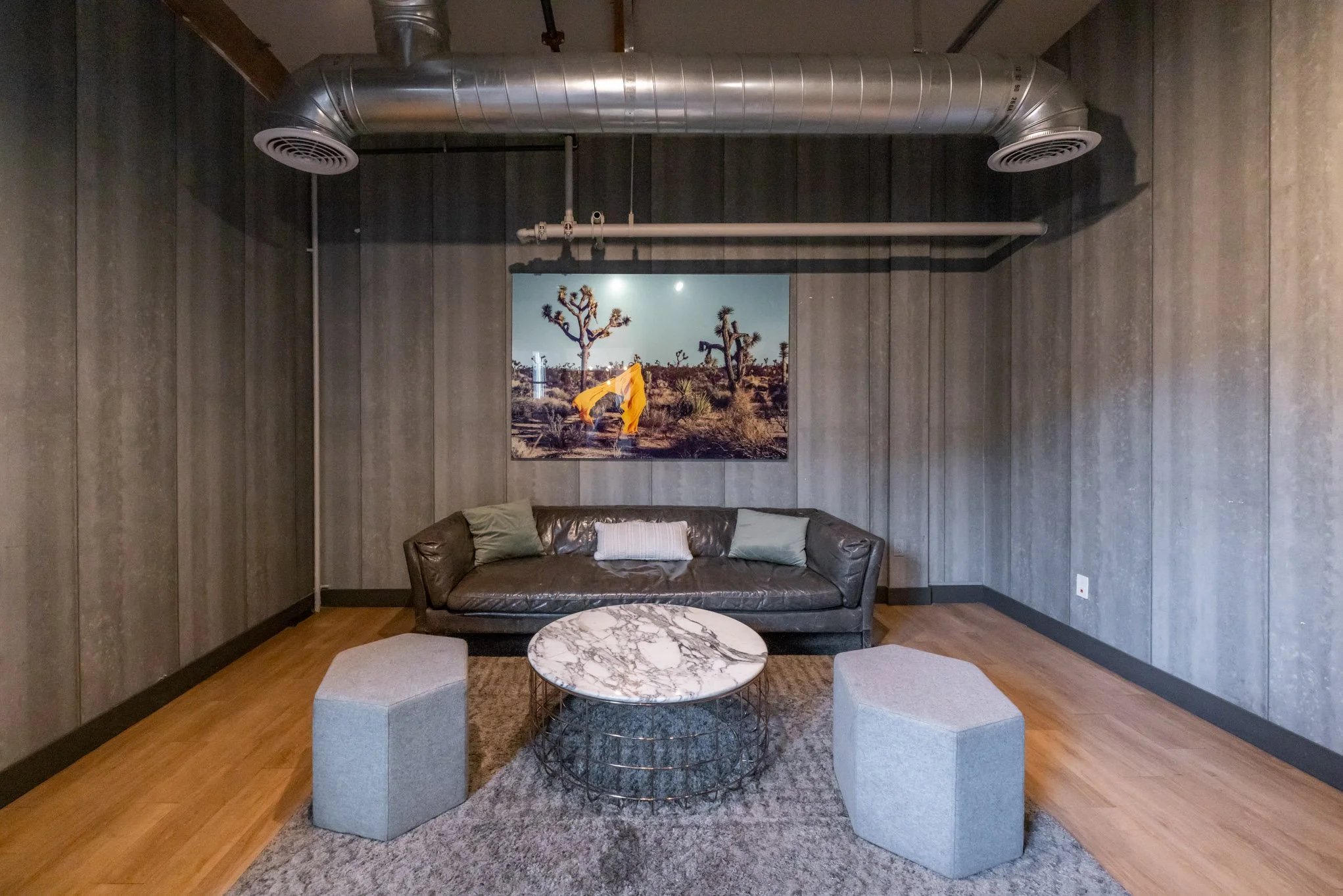 Modern living room with a black leather couch, a marble-topped coffee table, two gray cube ottomans, a painting of desert landscape with Joshua trees, and industrial-style exposed ductwork on a gray wood-paneled wall.