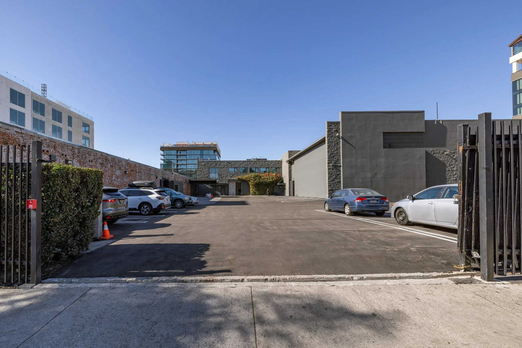 Parking lot with several parked cars and tall modern buildings in the background under a clear blue sky.