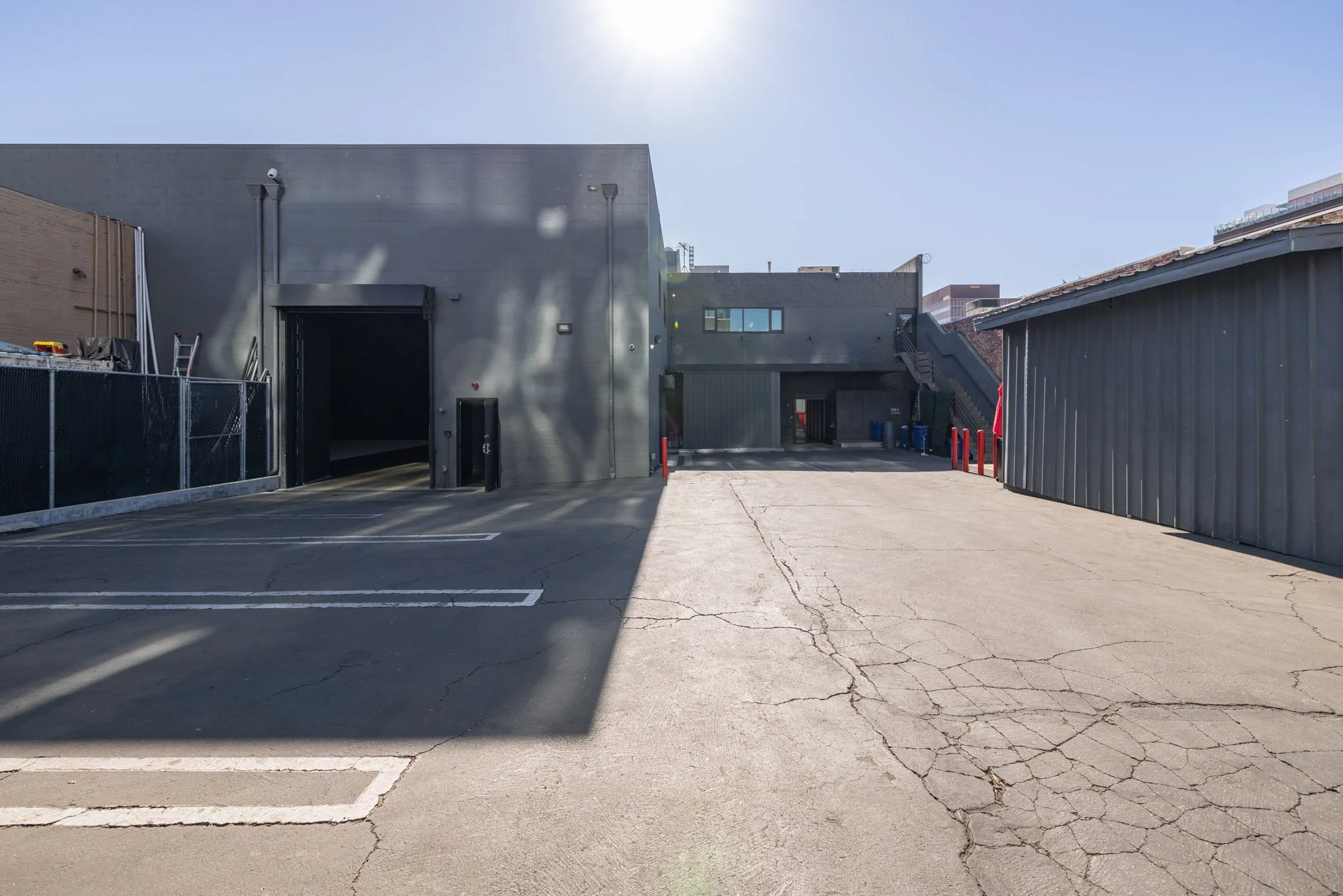 Empty parking lot with cracked pavement, adjacent gray Industrial building on right, smooth dark building with stairway in the background, bright sunny day.