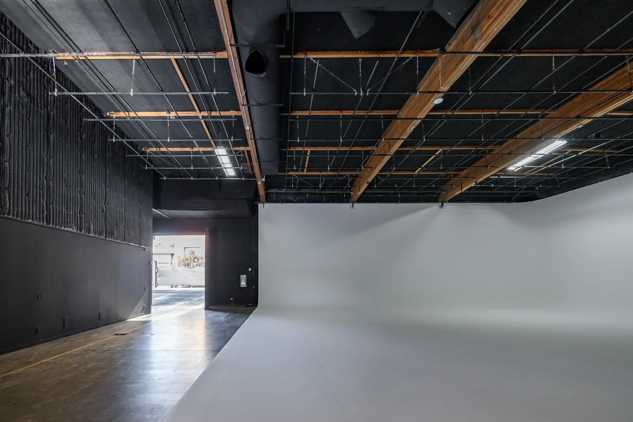 Empty photo studio with a white backdrop and black walls, partially visible. The ceiling has exposed black and wooden beams, pipes, and lighting fixtures. The floor is concrete, and there's an open door at the back allowing natural light.