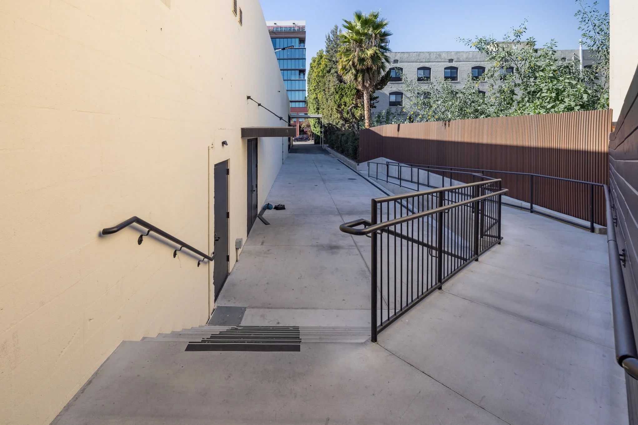 A sidewalk and stairs with black handrails next to a beige building with two black doors, a brown wooden fence, and trees in an urban setting with buildings in the background.