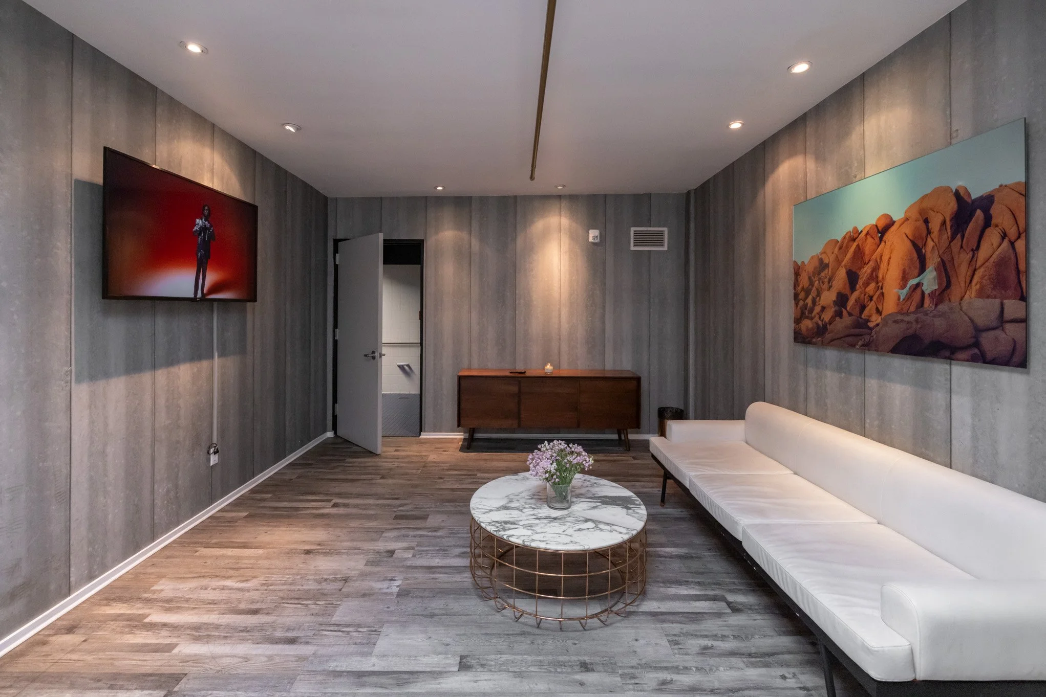 Modern waiting room with gray wood-paneled walls, a white sofa, a round marble coffee table with flowers, a wooden sideboard, a flat-screen TV, and framed landscape art.