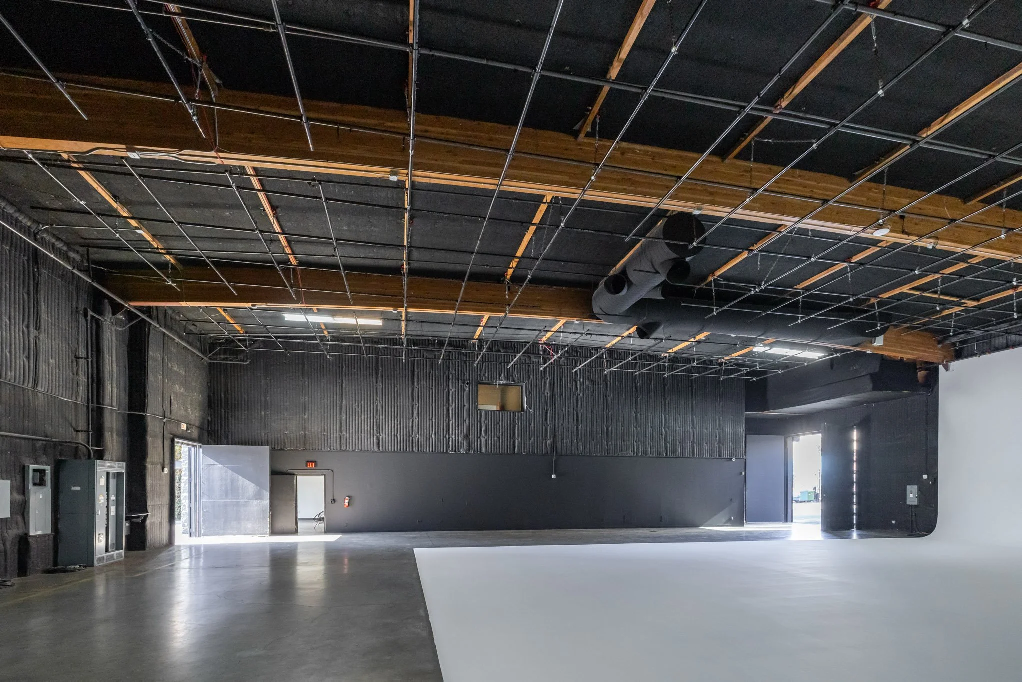 Empty industrial style interior with high ceilings, exposed ductwork, black walls, black ceiling grid, and a large bright open door at the back.