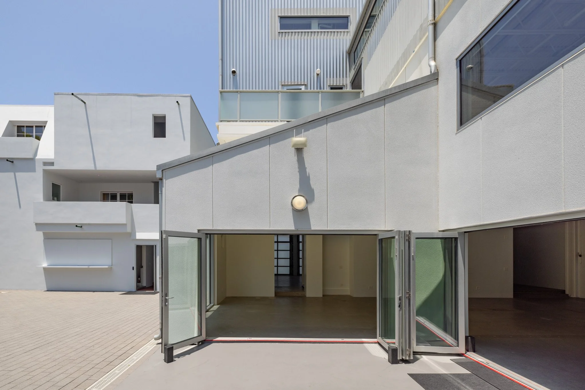 Modern white building with open glass doors leading to an empty indoor space, outdoor paved area, and neighboring buildings with balconies.