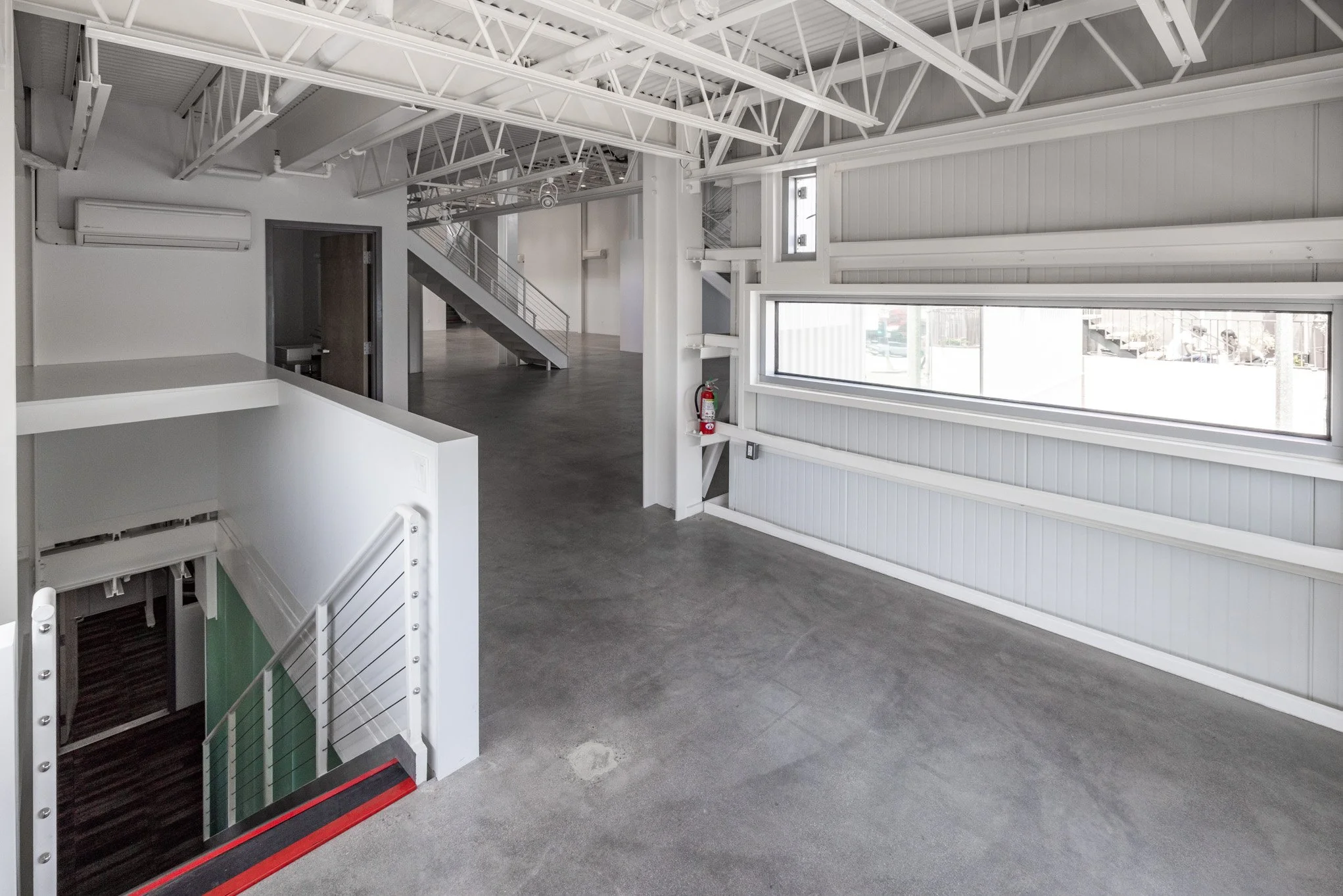 Empty interior space with white painted metal beams, large rectangular window, gray floors, and a staircase leading to an upper level.