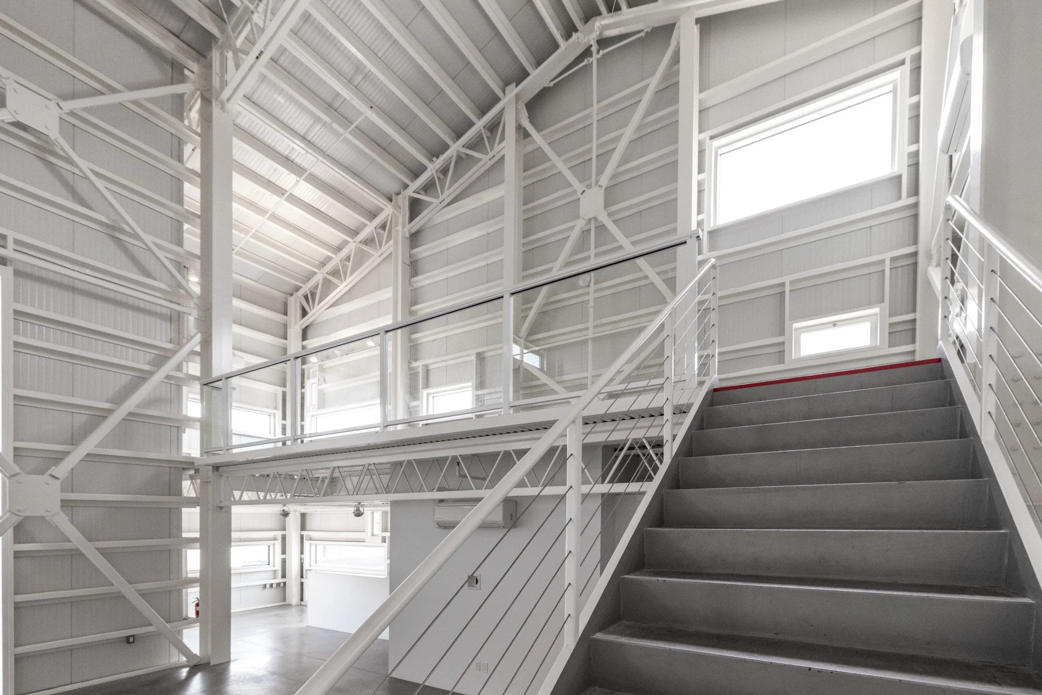 Interior of a modern, industrial-style building with white metal walls, multiple windows, and a staircase made of gray concrete steps with white metal railings.