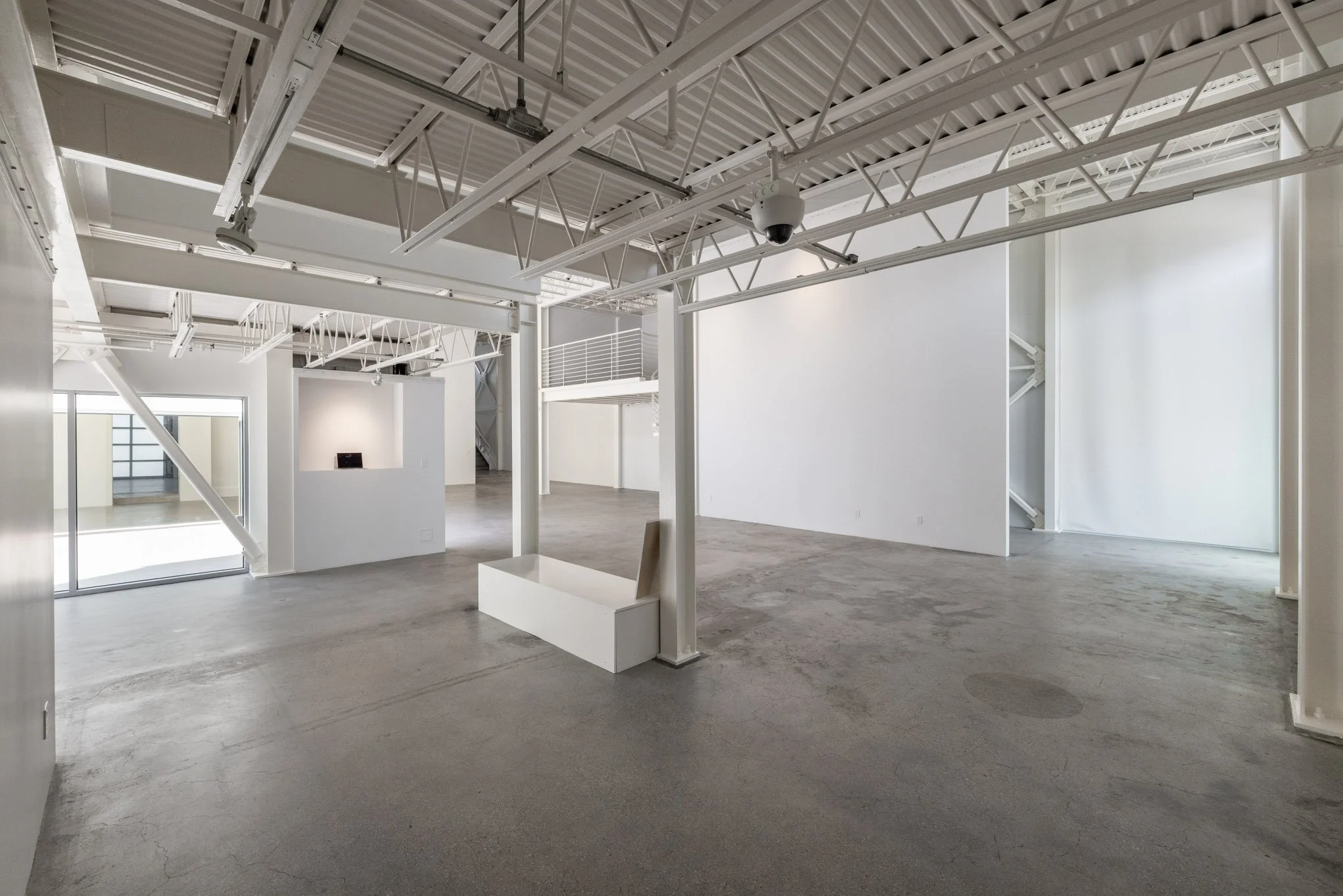 Empty, modern gallery or art space with white walls, concrete floors, and exposed ceiling with lighting fixtures and security cameras.