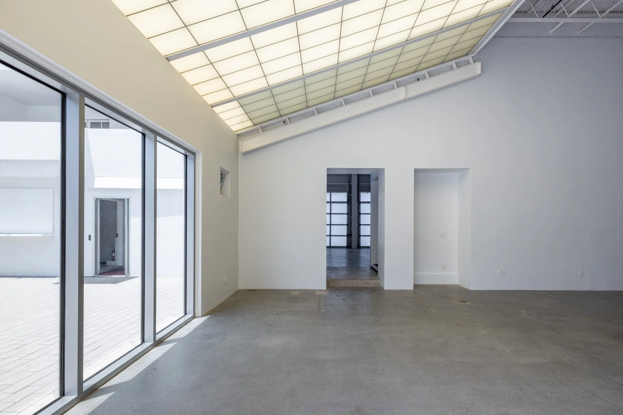 Empty room with large glass sliding doors, white walls, high ceiling with glow panels, and a concrete floor.