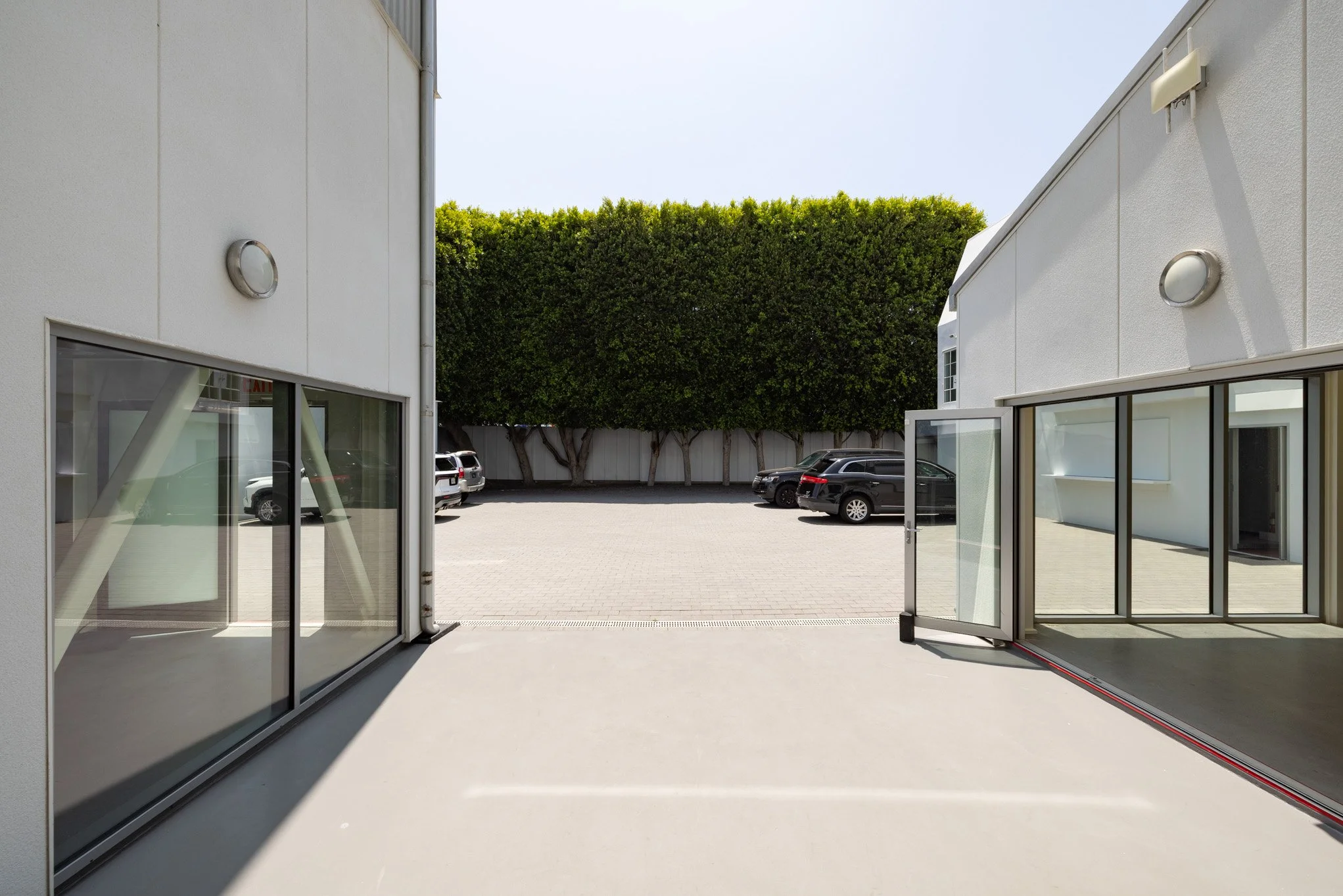 View of a parking lot with several cars, seen from inside a modern building with glass sliding doors and exterior white walls. There is a row of tall green trees at the back of the parking lot.
