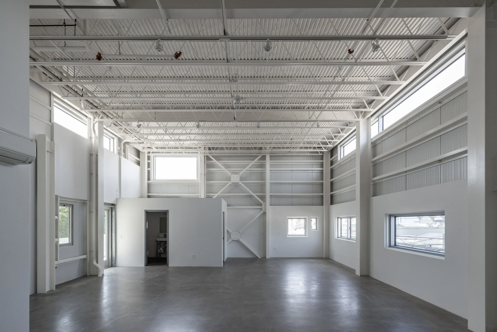Empty industrial or warehouse space with white walls, high ceiling, metal beams, and multiple windows allowing natural light.