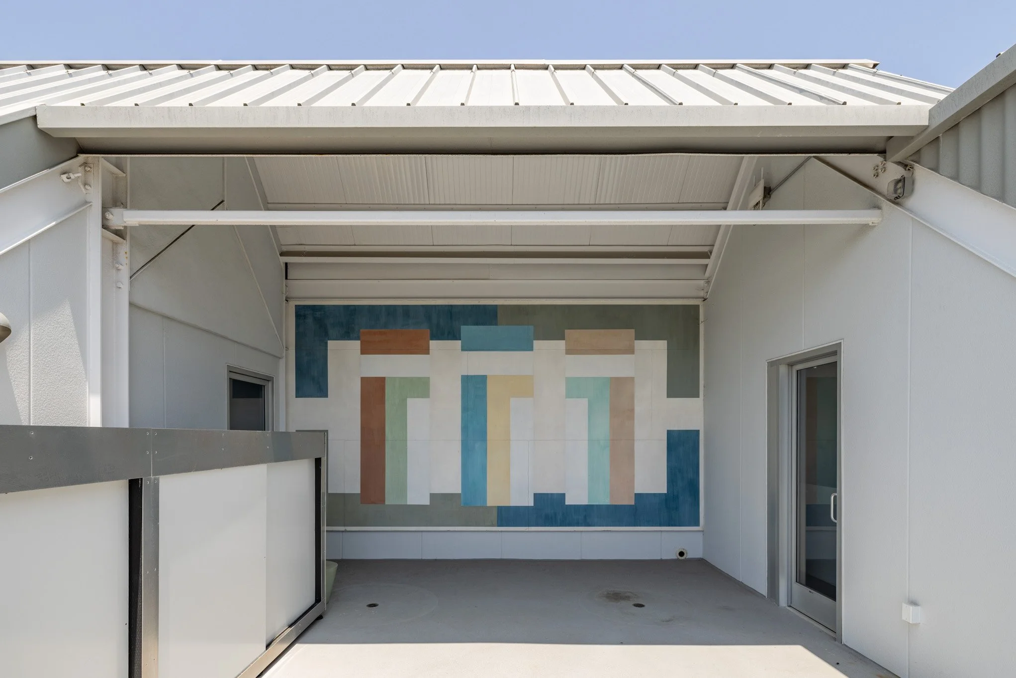 A covered balcony with a colorful geometric mural on the back wall, white walls, and a sliding glass door.