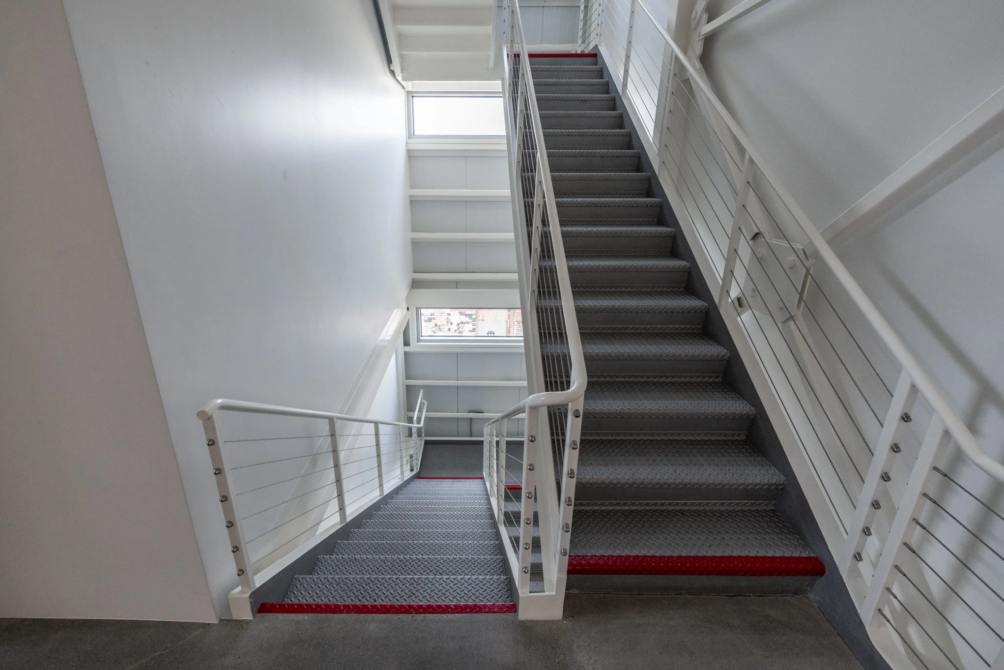 Indoor staircase with metal steps and white railings in a well-lit building, with large windows at the top.