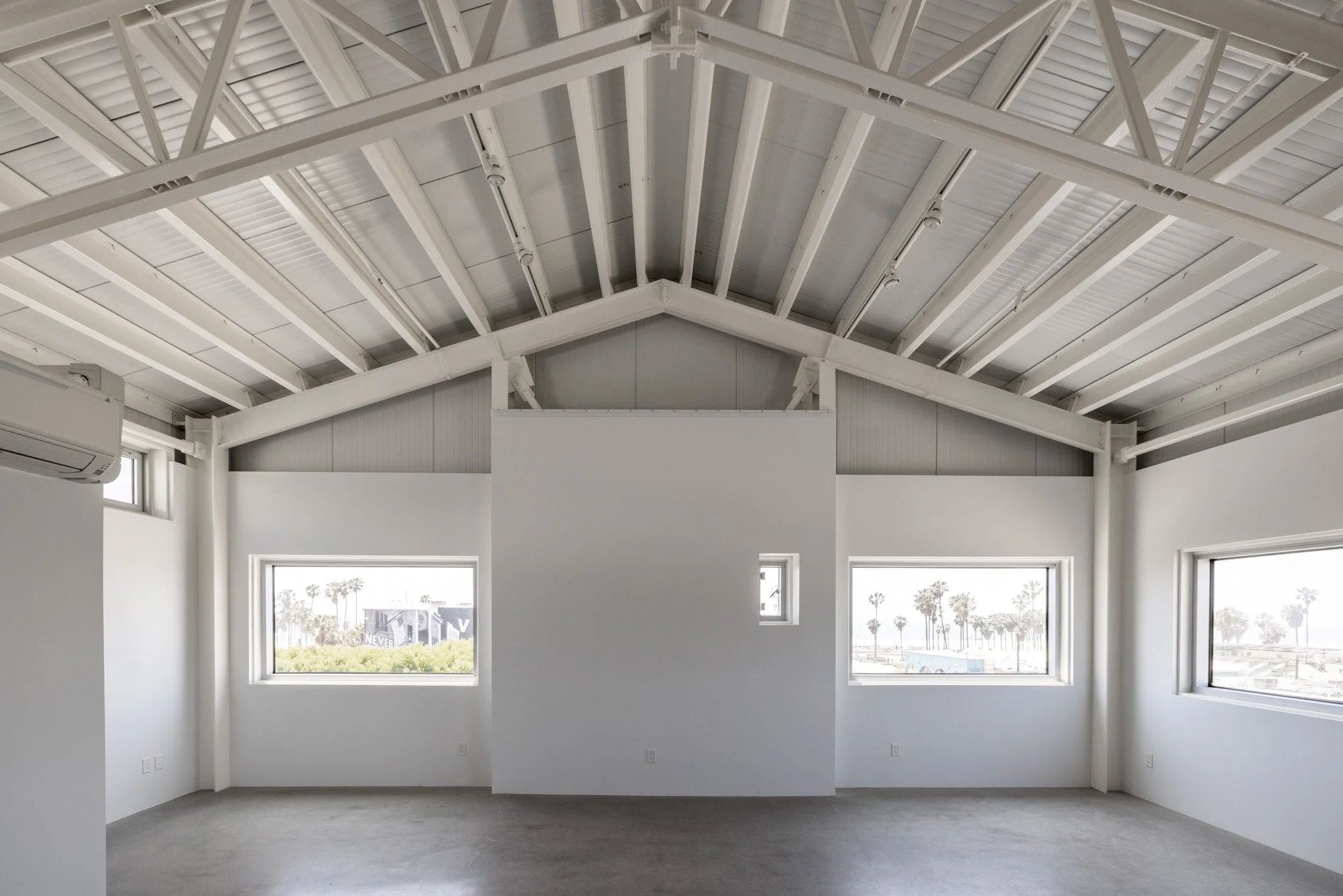 Interior of an unfinished room with white walls, windows, an exposed ceiling with metal beams, and skylights, overlooking a sunny outdoor scene with palm trees.