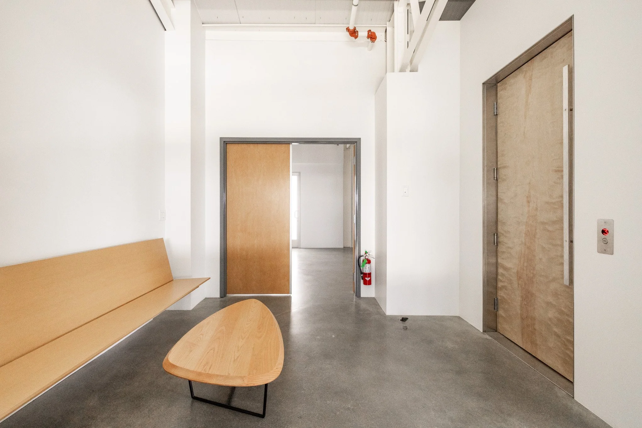 Modern, minimalistic waiting area with wooden bench, small wooden table with black metal legs, white walls, concrete floor, and an elevator with wooden doors. Fire extinguisher is on the wall near doorway, and there is an unseen area beyond the slidi