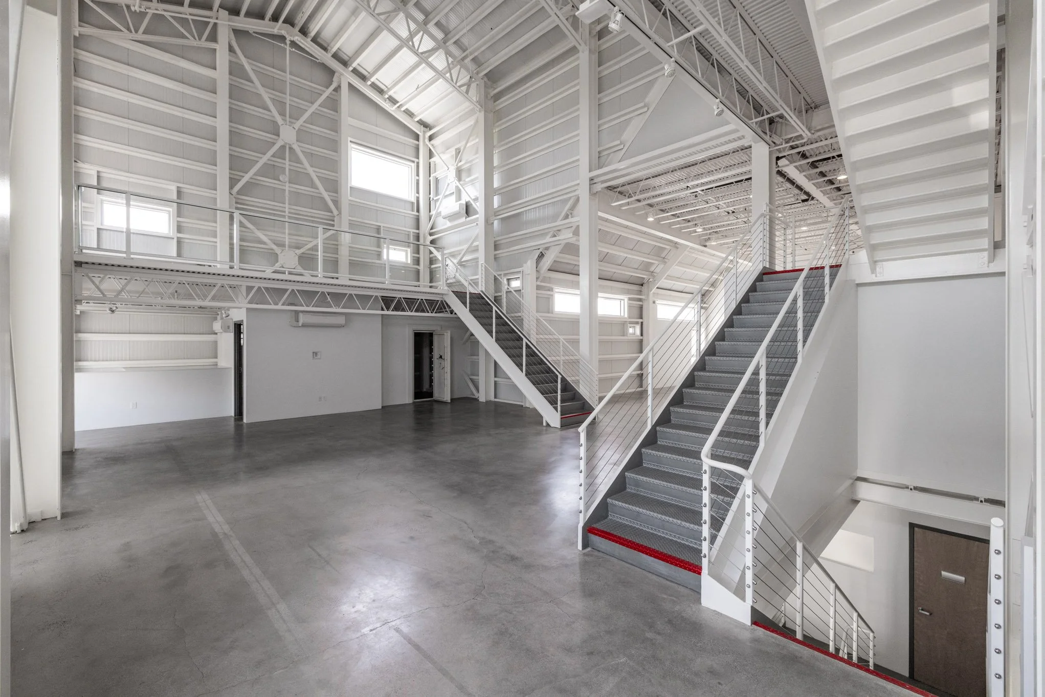 Empty industrial-style interior space with steel staircase and mezzanine, concrete floor, white walls, and large windows.