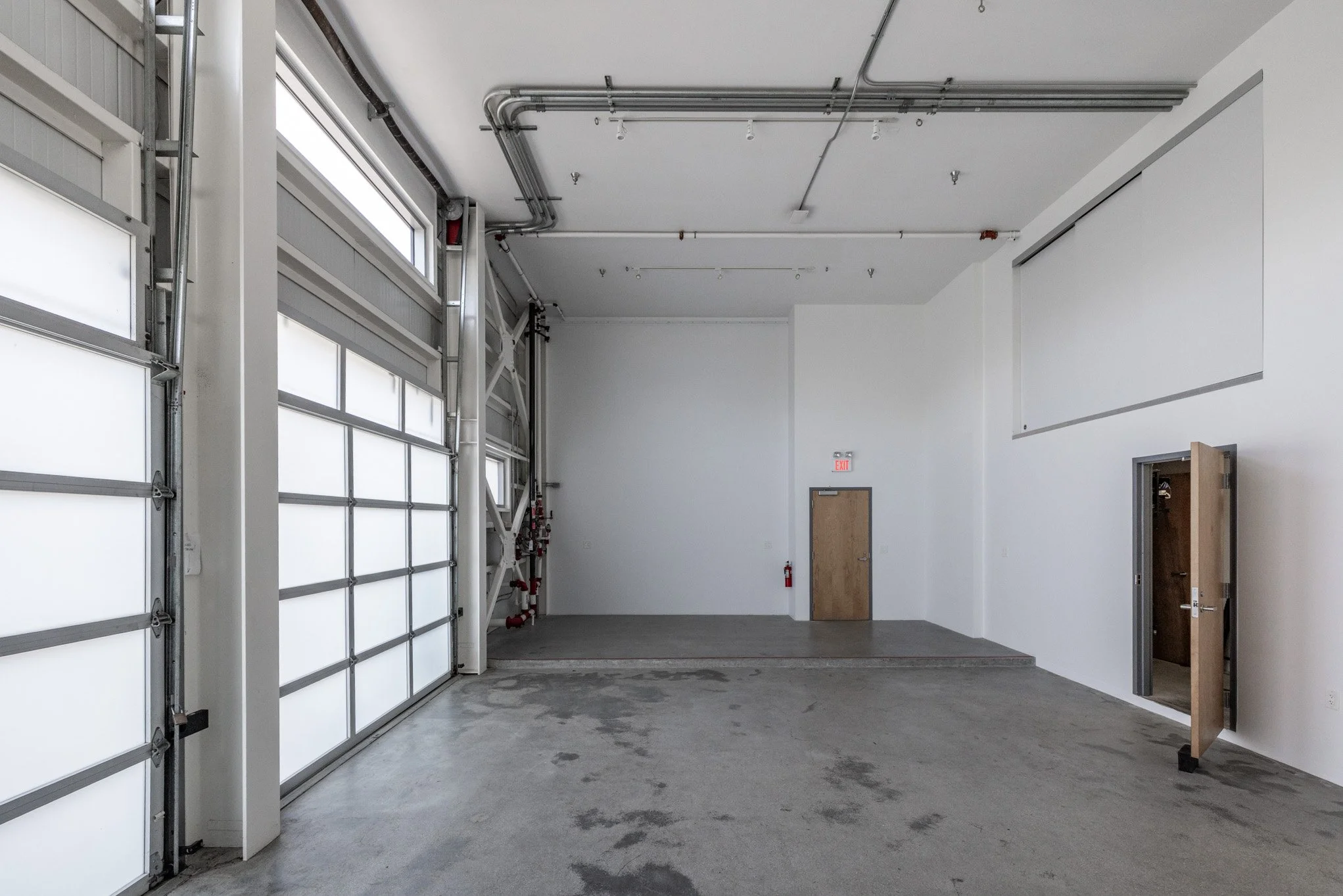 Empty industrial garage or warehouse with large white garage doors on the left, a small door on the back wall, and an open door on the right wall. The floors are concrete, and the walls are white.