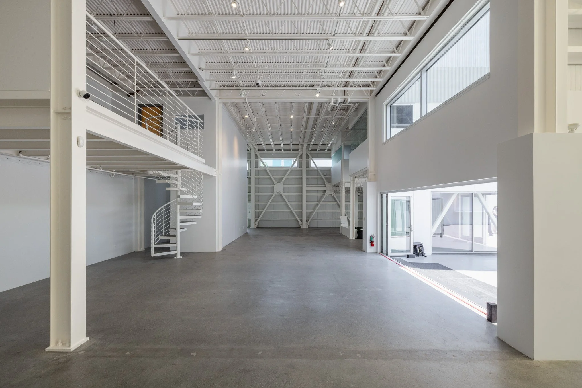 Empty industrial or commercial warehouse interior with high ceiling, large windows, white metal spiral staircase, and concrete floor.