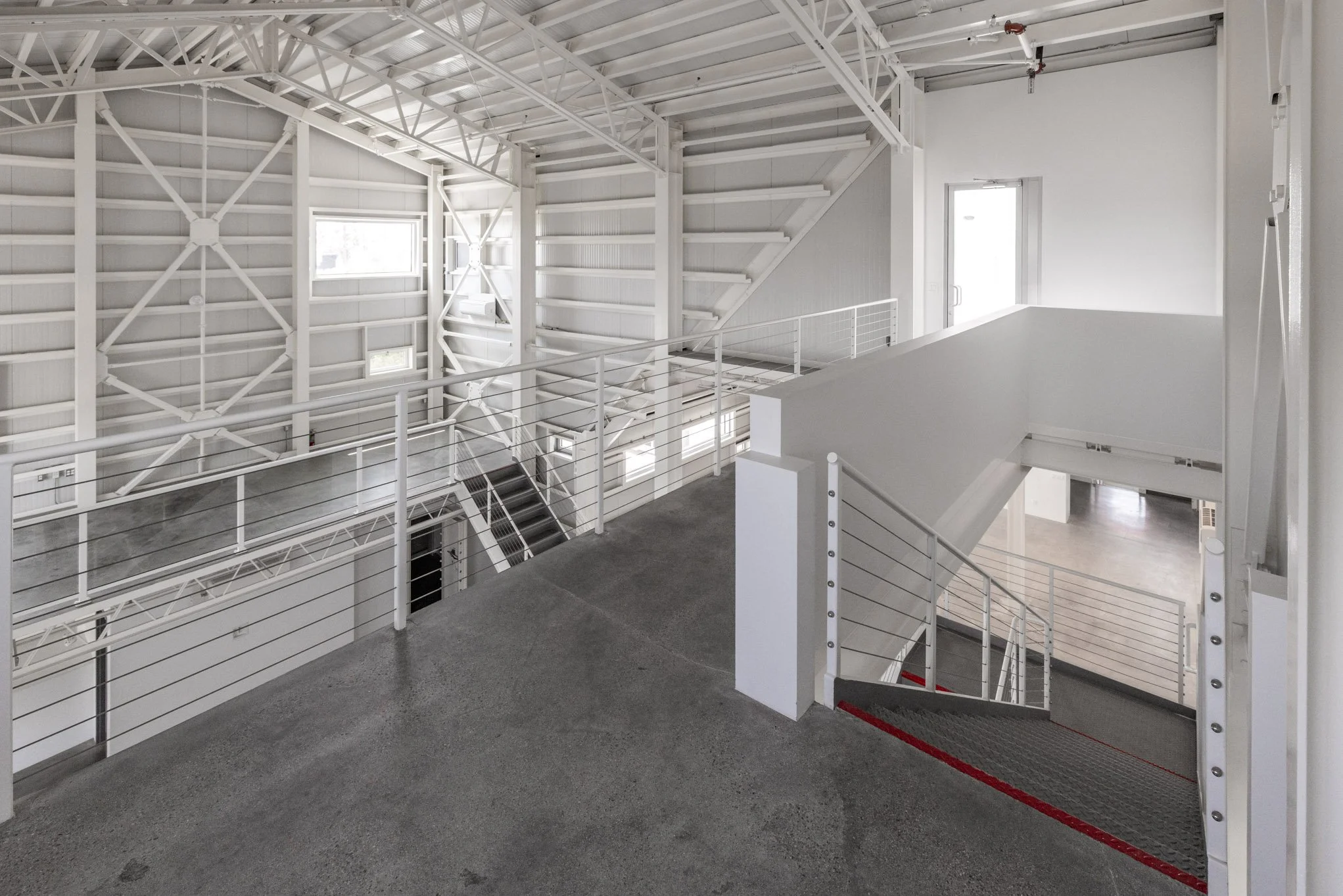 Interior of a modern, industrial-style building with white metal beams, white walls, and gray concrete floors, featuring multiple staircases and railings.