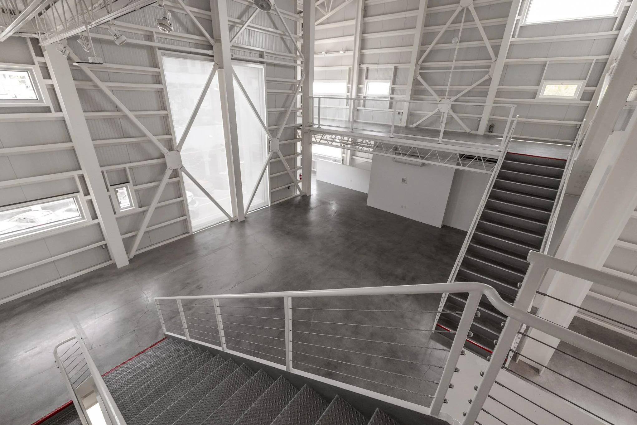 Empty modern industrial-style interior with metal stairs, white walls, large windows, and a concrete floor.