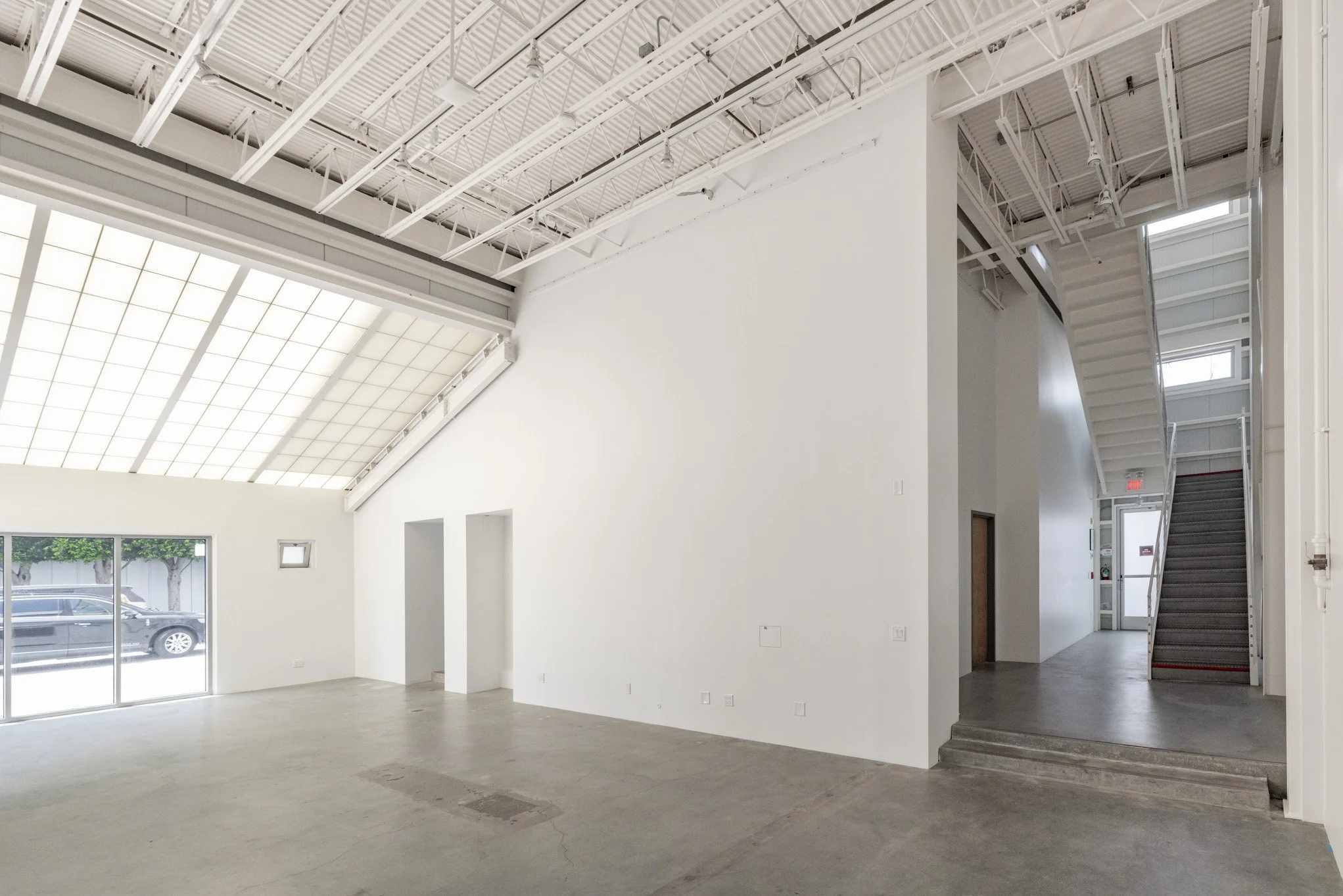 Empty interior space with white walls, high ceiling with exposed beams, large window and a staircase leading to an upper level.